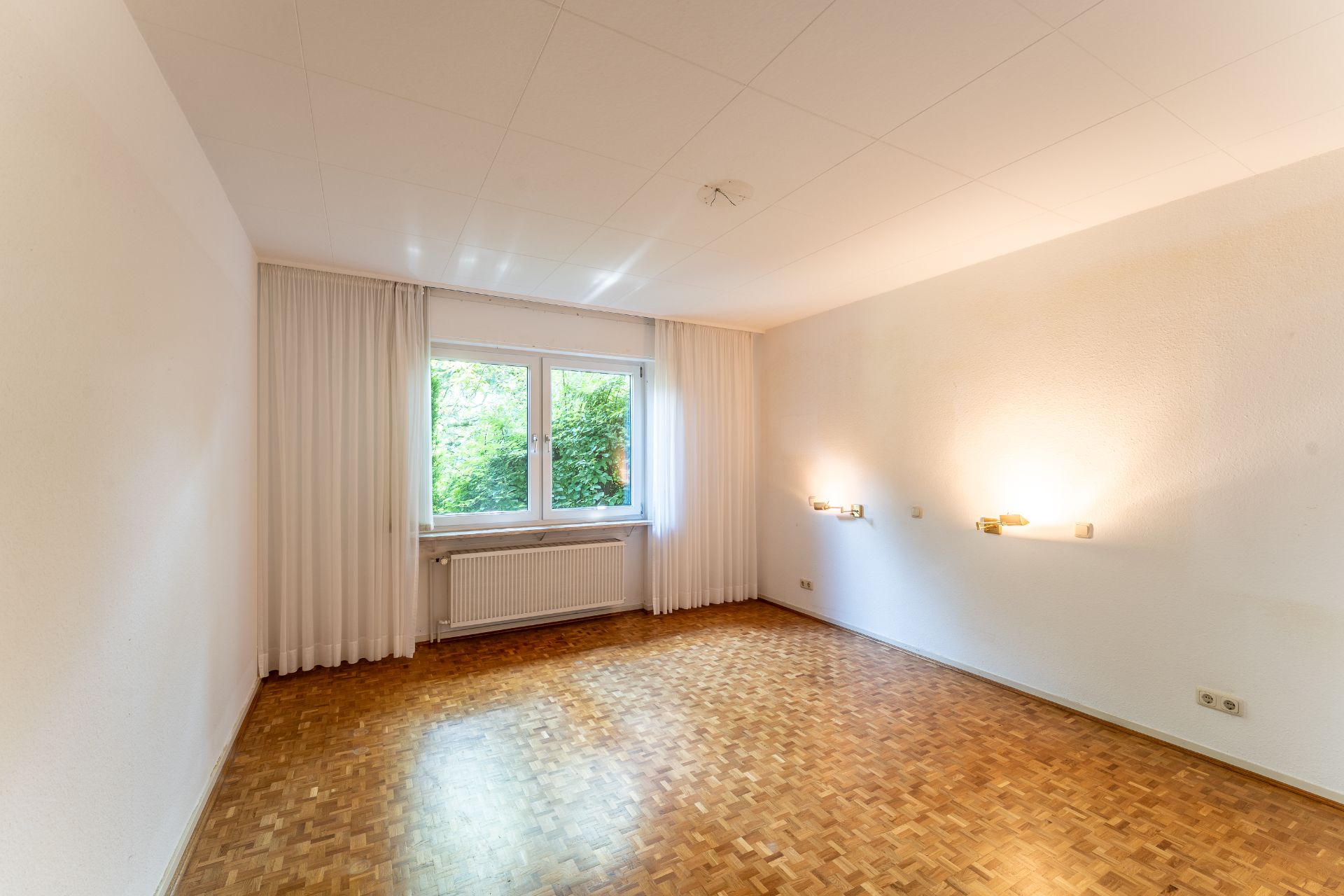 Leerer Raum mit Parkettboden, Fenster mit Vorhängen und zwei Wandleuchten.