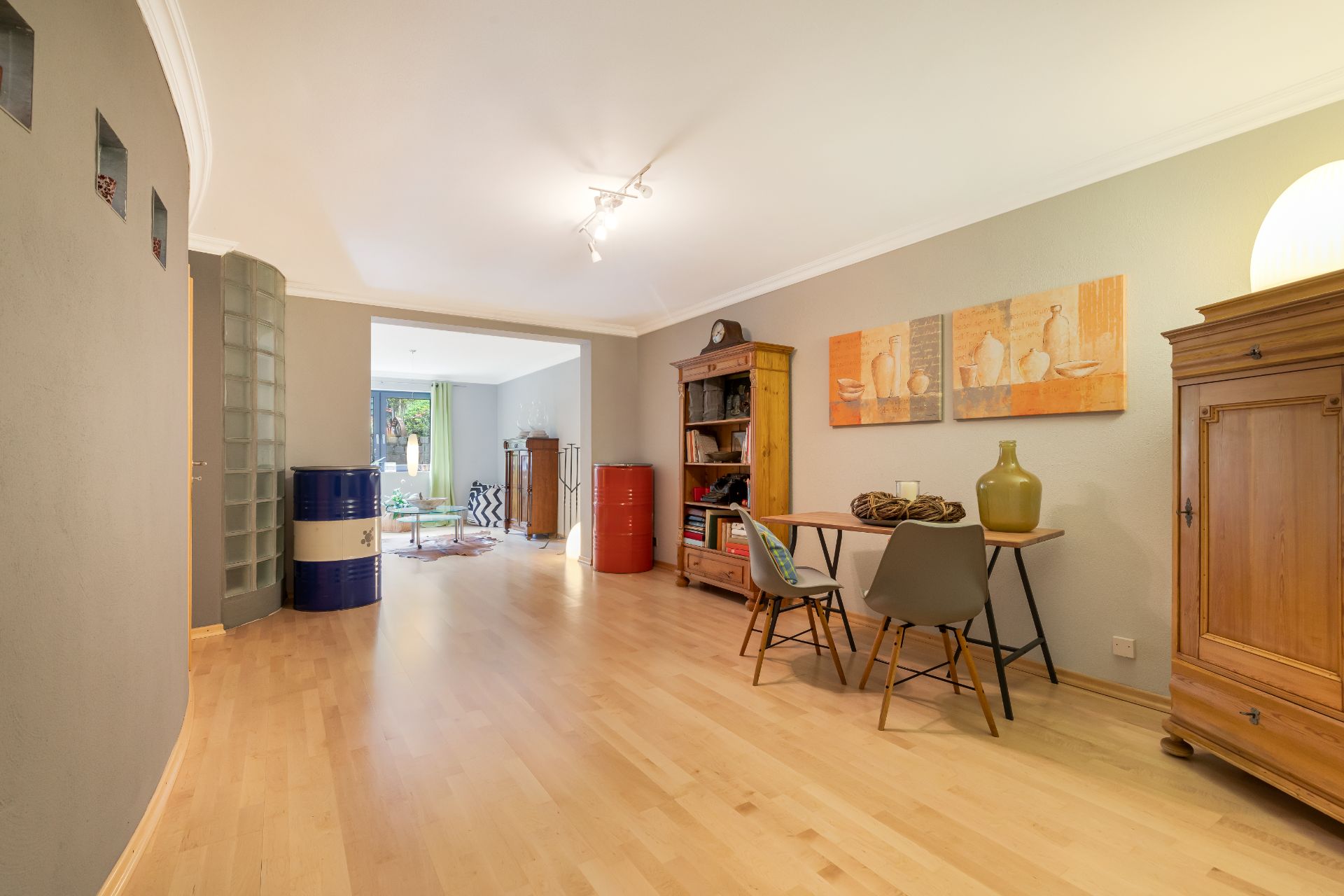 Heller Raum mit Holzboden, Schreibtisch, Stühlen, Regal, Kunst an der Wand und Zugang zu weiterem Zimmer.