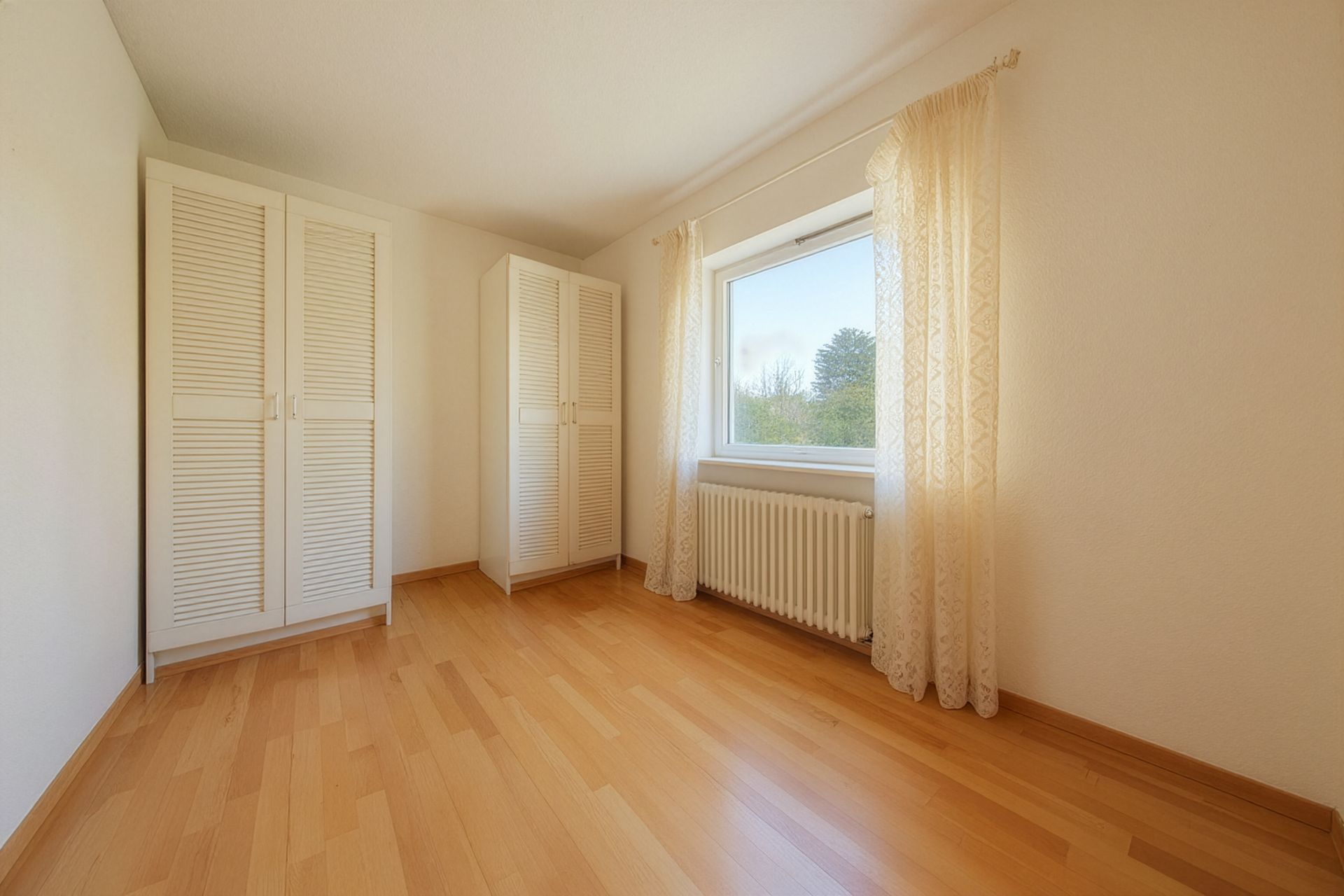 Helles Zimmer mit Holzfußboden, zwei weißen Schränken und einem Fenster mit Gardinen.