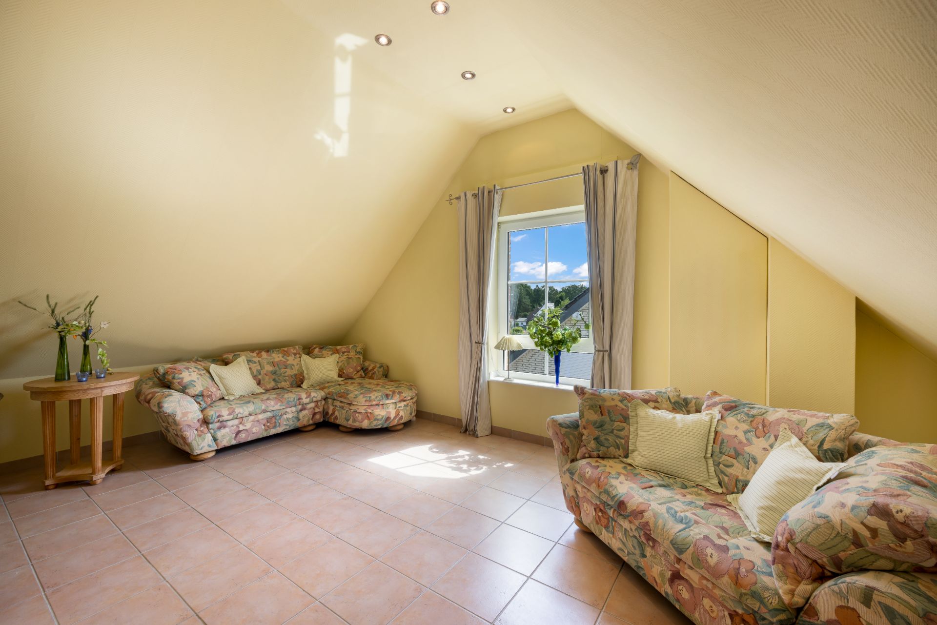 Helles Wohnzimmer mit zwei gemusterten Sofas, einem Fenster und einem Beistelltisch mit Blumen.
