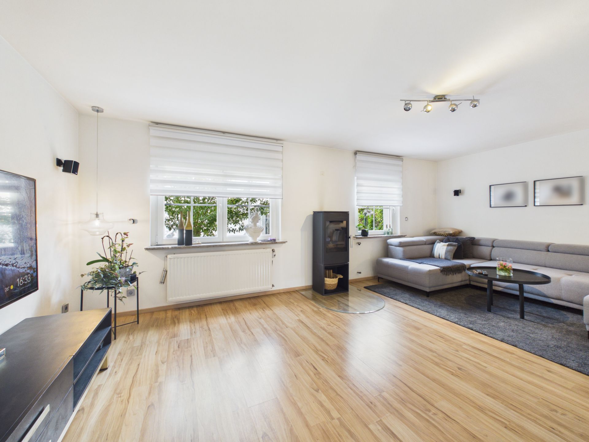 Helles Wohnzimmer mit Ecksofa, Holzfußboden, Kaminofen und zwei großen Fenstern.