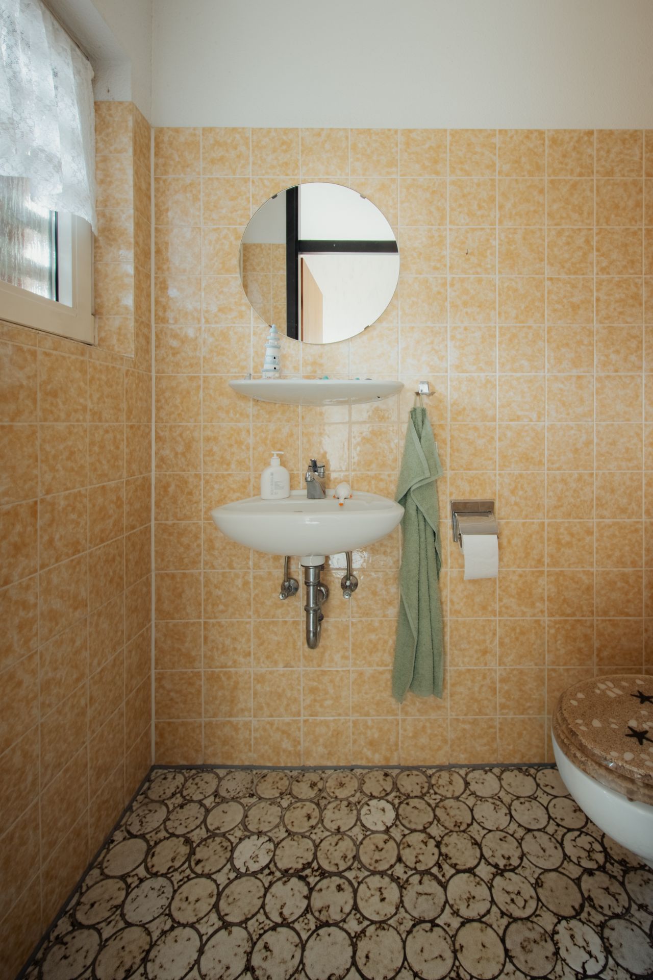Kleines Badezimmer mit Waschbecken, rundem Spiegel, Handtuch und Toilettenpapierhalter.