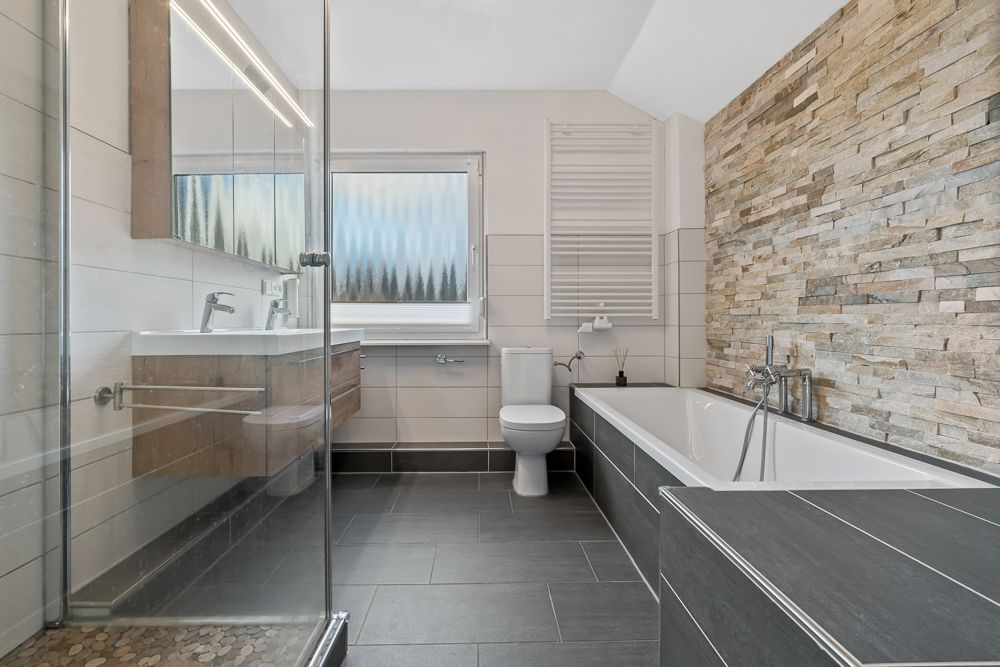 Modernes Badezimmer mit Dusche, Doppelwaschbecken, Toilette und Badewanne, Steinwand rechts.