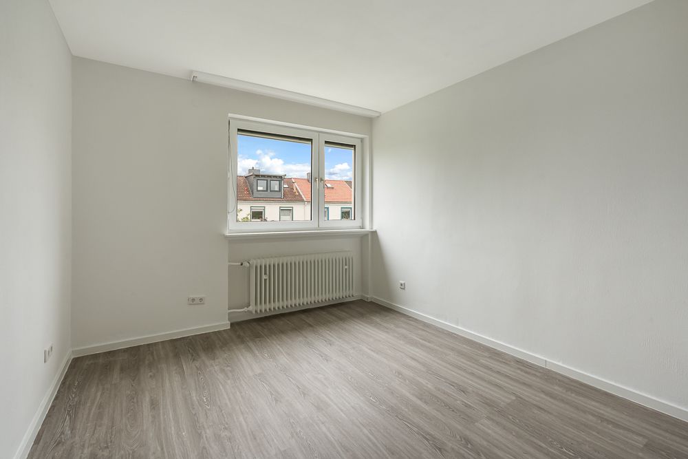Leeres Zimmer mit grauem Boden, weißer Wand und Fenster mit Blick auf Nachbarhäuser.