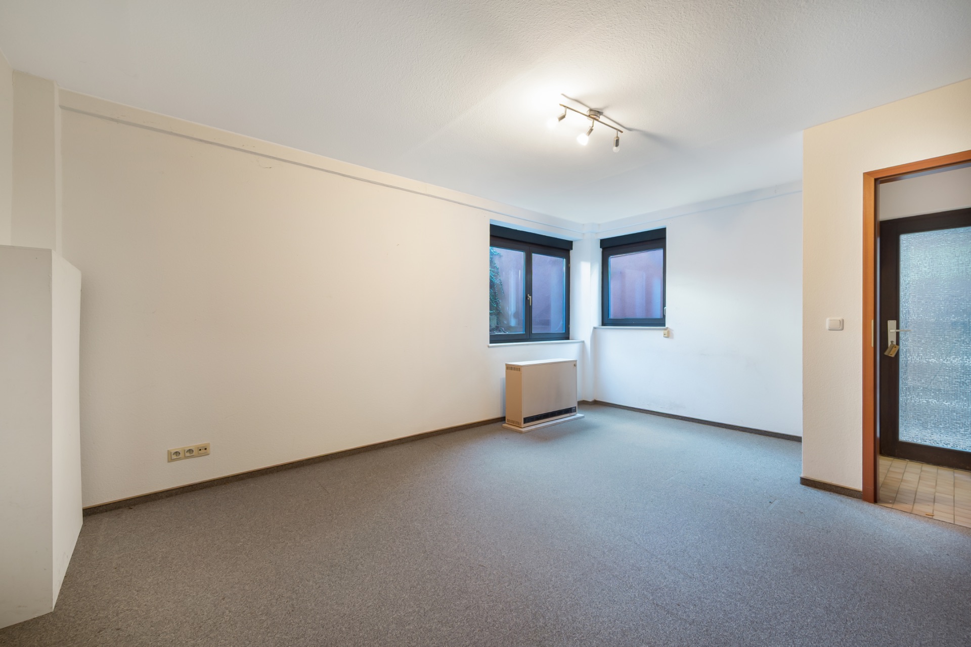 Helles, leeres Zimmer mit zwei Fenstern, Teppichboden und Deckenleuchte.
