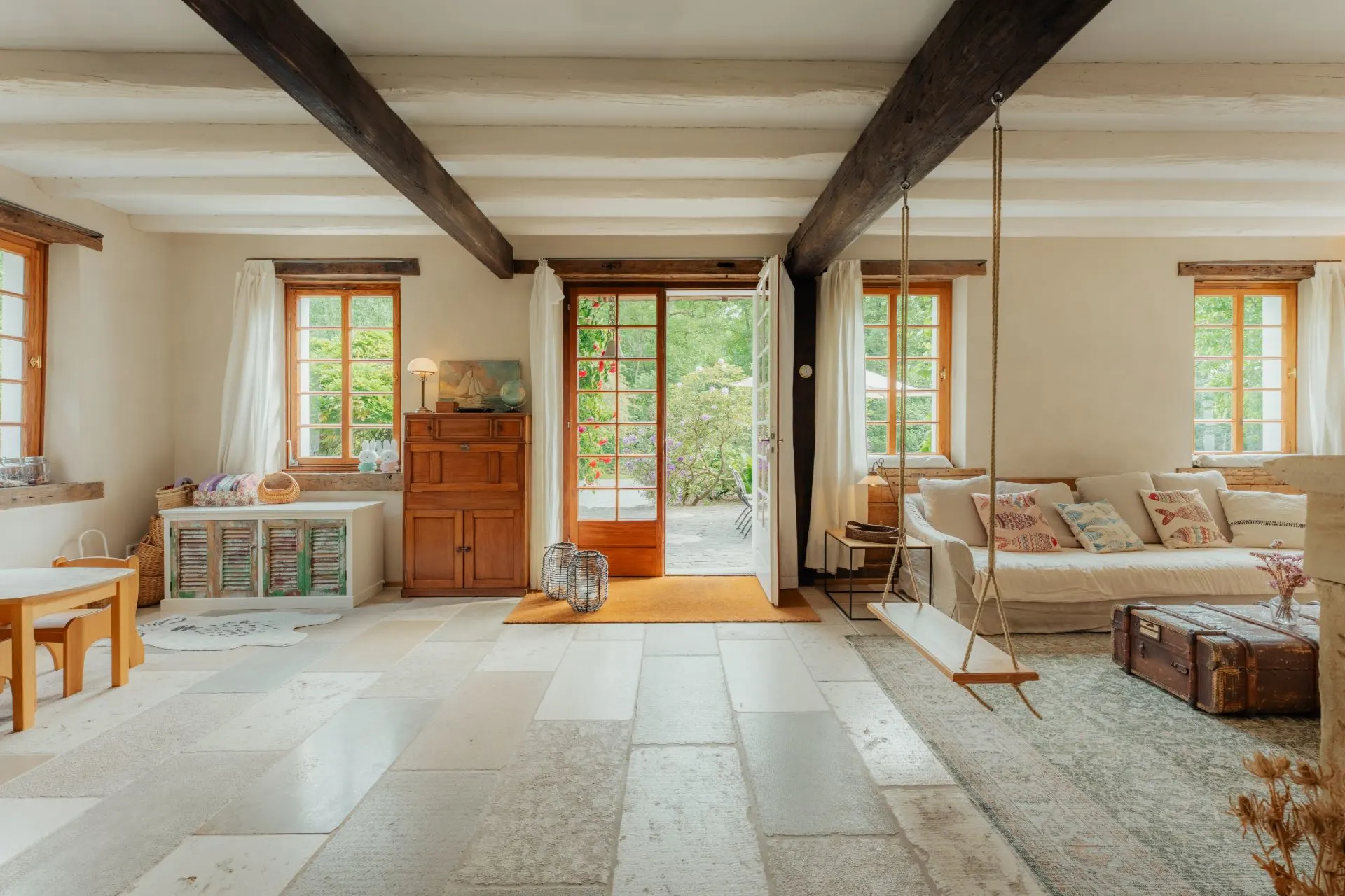 Helles Wohnzimmer mit Schaukel, Sofa, Holzbalken, großen Fenstern und offenem Zugang zum Garten.