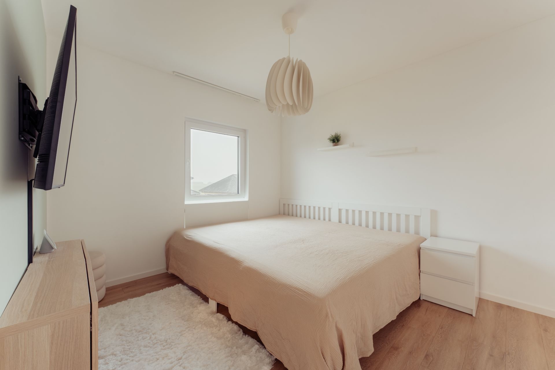 Helles Schlafzimmer mit Doppelbett, Nachttisch, Teppich, Fernseher und Fenster.