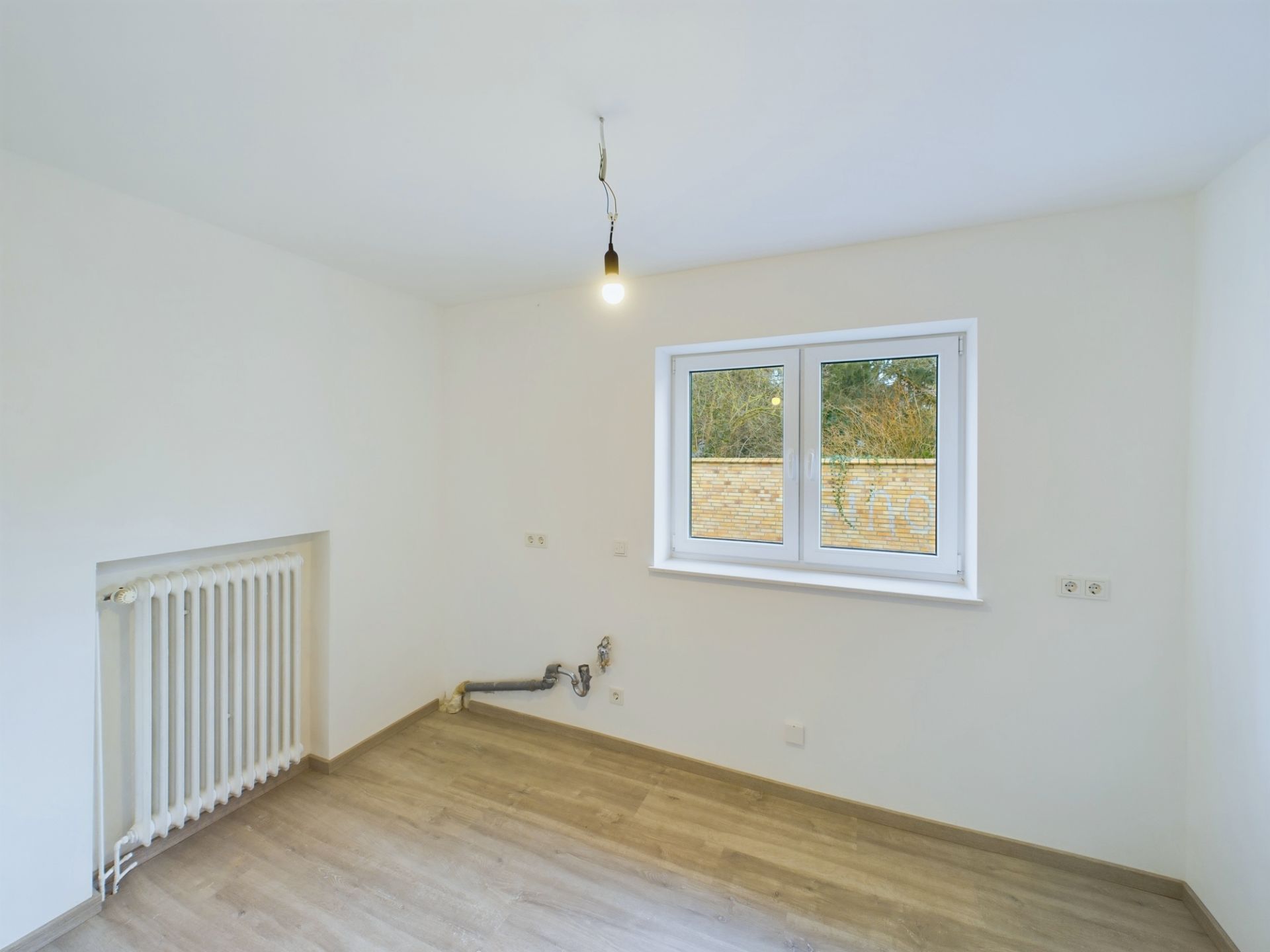 Leerer Raum mit Fenster, Heizung, hellen Wänden und Anschlüssen an der Wand.