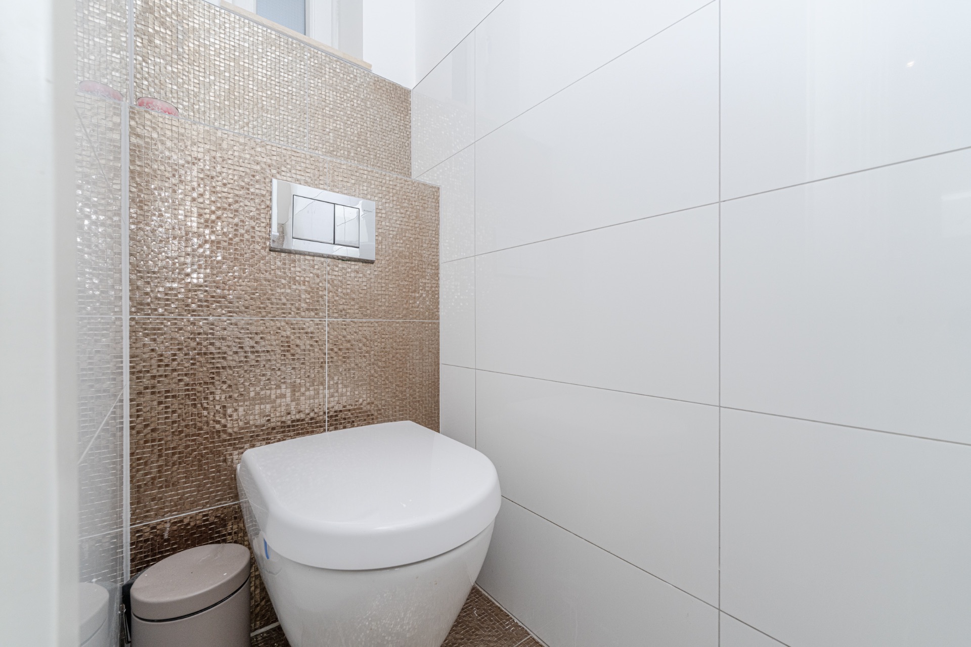 Modernes WC mit weißen Fliesen und goldener Mosaikwand, kleiner Abfalleimer daneben.
