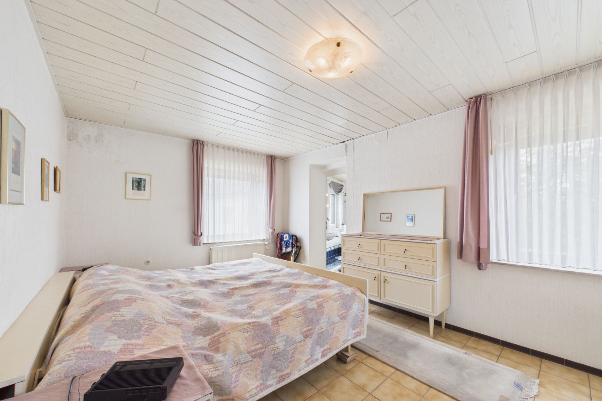 Helles Schlafzimmer mit Doppelbett, Kommode und zwei Fenstern mit Vorhängen.