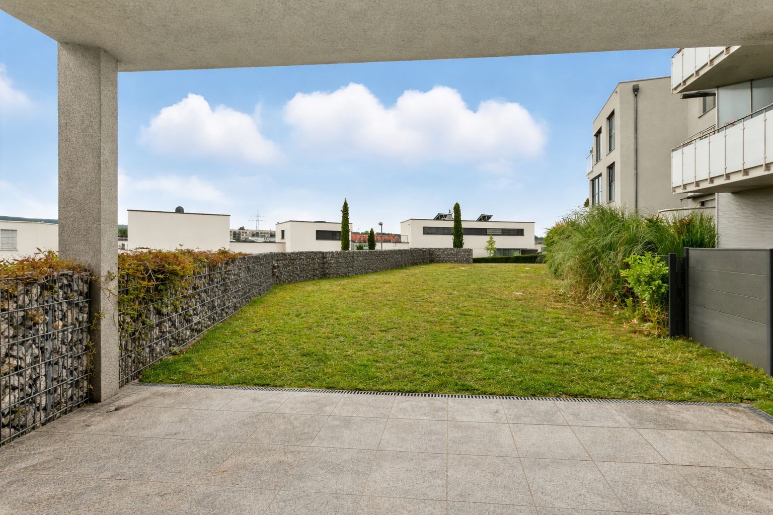 Blick von einer überdachten Terrasse auf einen gepflegten Rasen im Hinterhof mit modernen Gebäuden.