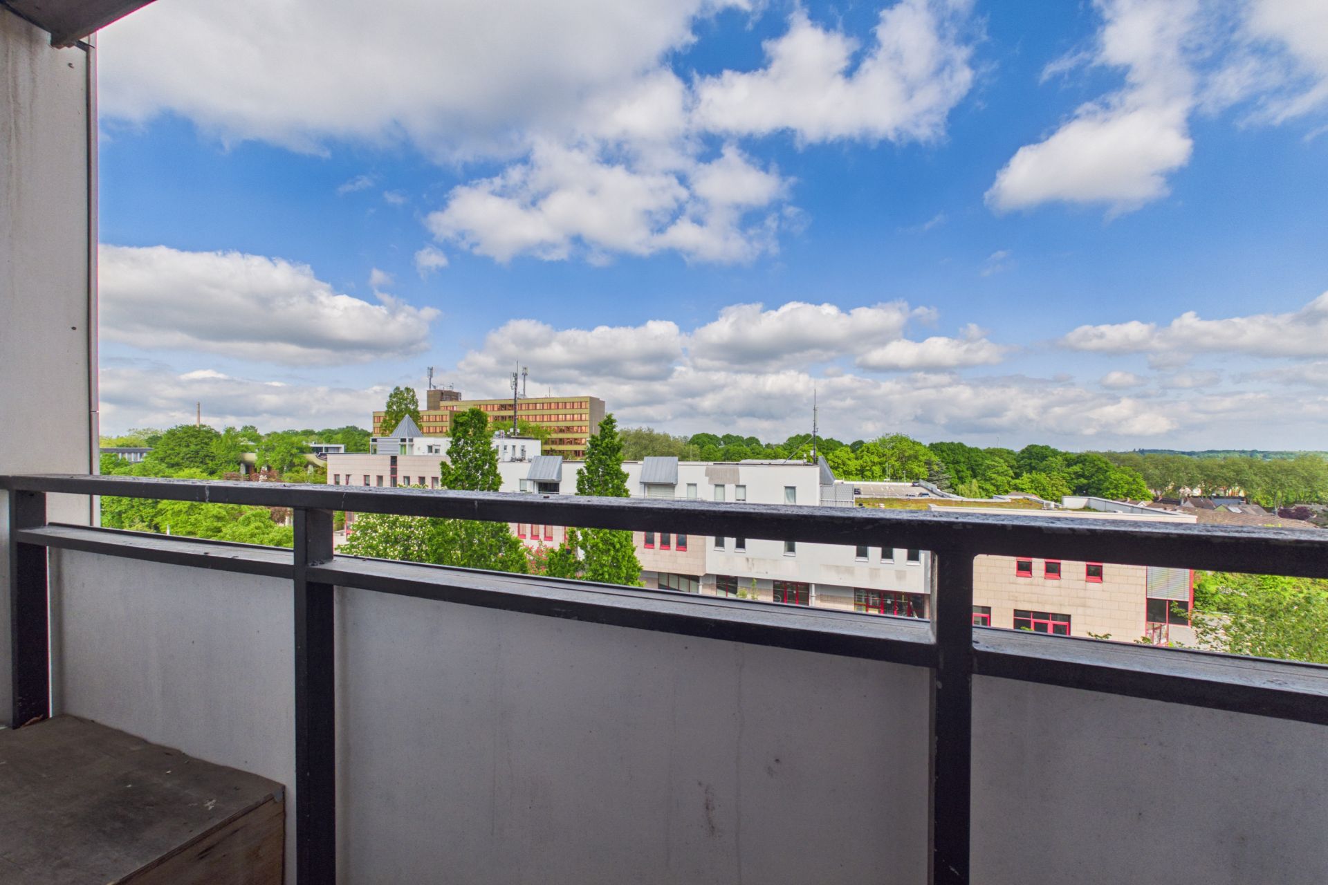 Blick von einem Balkon auf grüne Bäume, Gebäude und einen blauen Himmel mit Wolken.