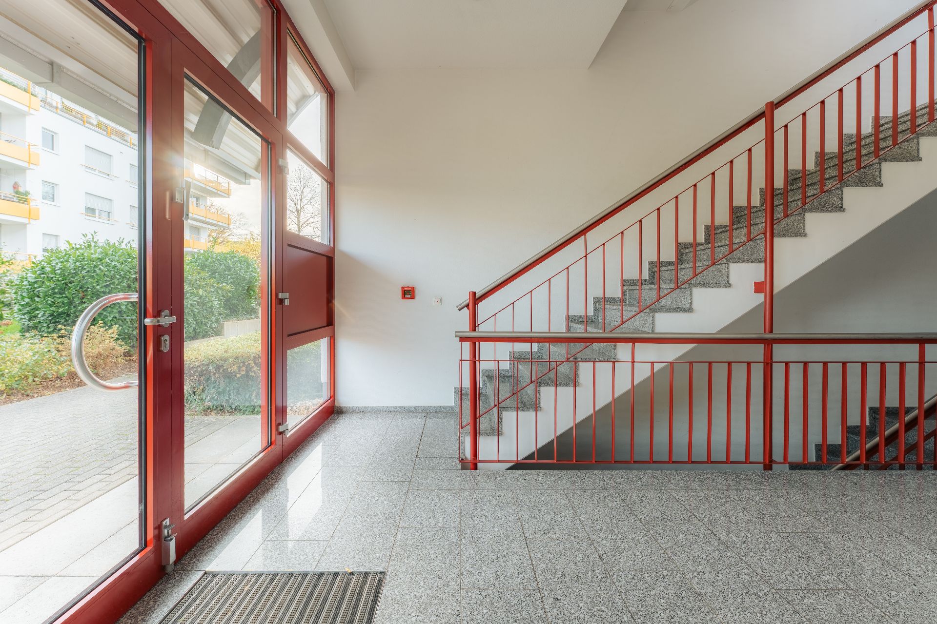 Heller Treppenhausbereich mit roten Geländern und Glastür, Blick nach draußen auf Grünfläche.