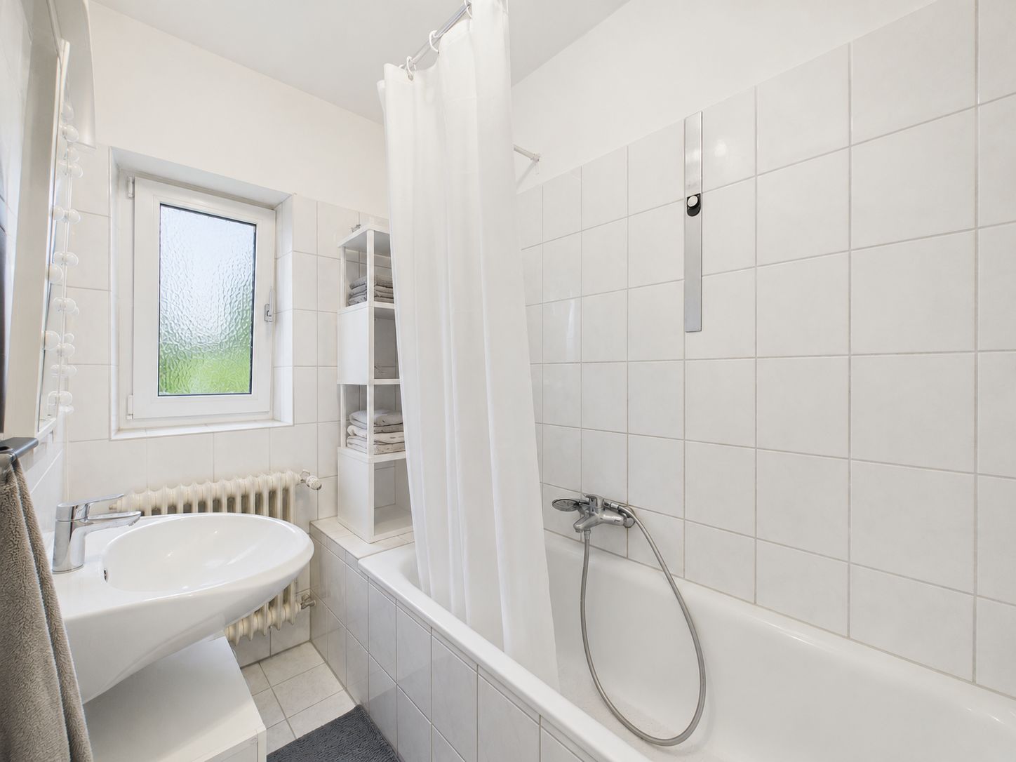 Helles Bad mit Badewanne, weißem Waschbecken, Fenster und Regal mit Handtüchern.