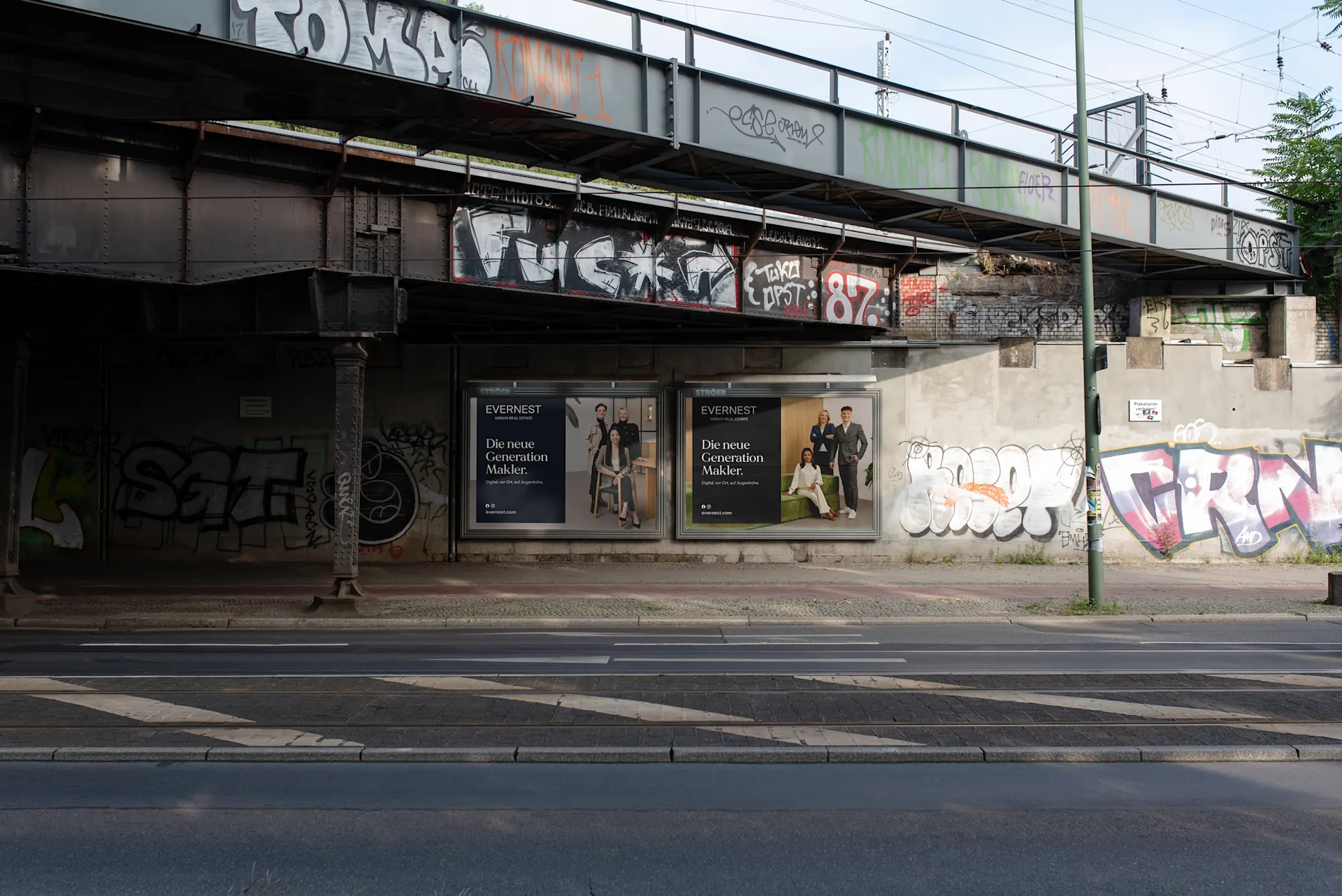Straße unter einer Bahnbrücke mit Graffiti und zwei Werbetafeln an der Wand.