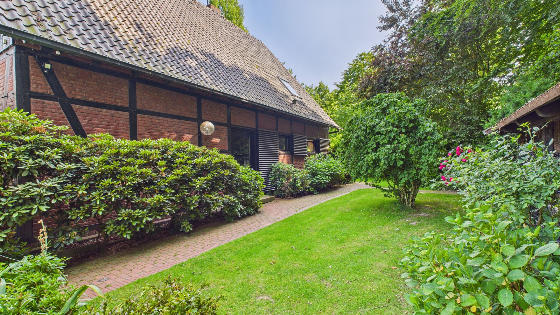 Gepflegter Garten mit Rasen und Büschen neben einem Backsteinhaus mit Satteldach.