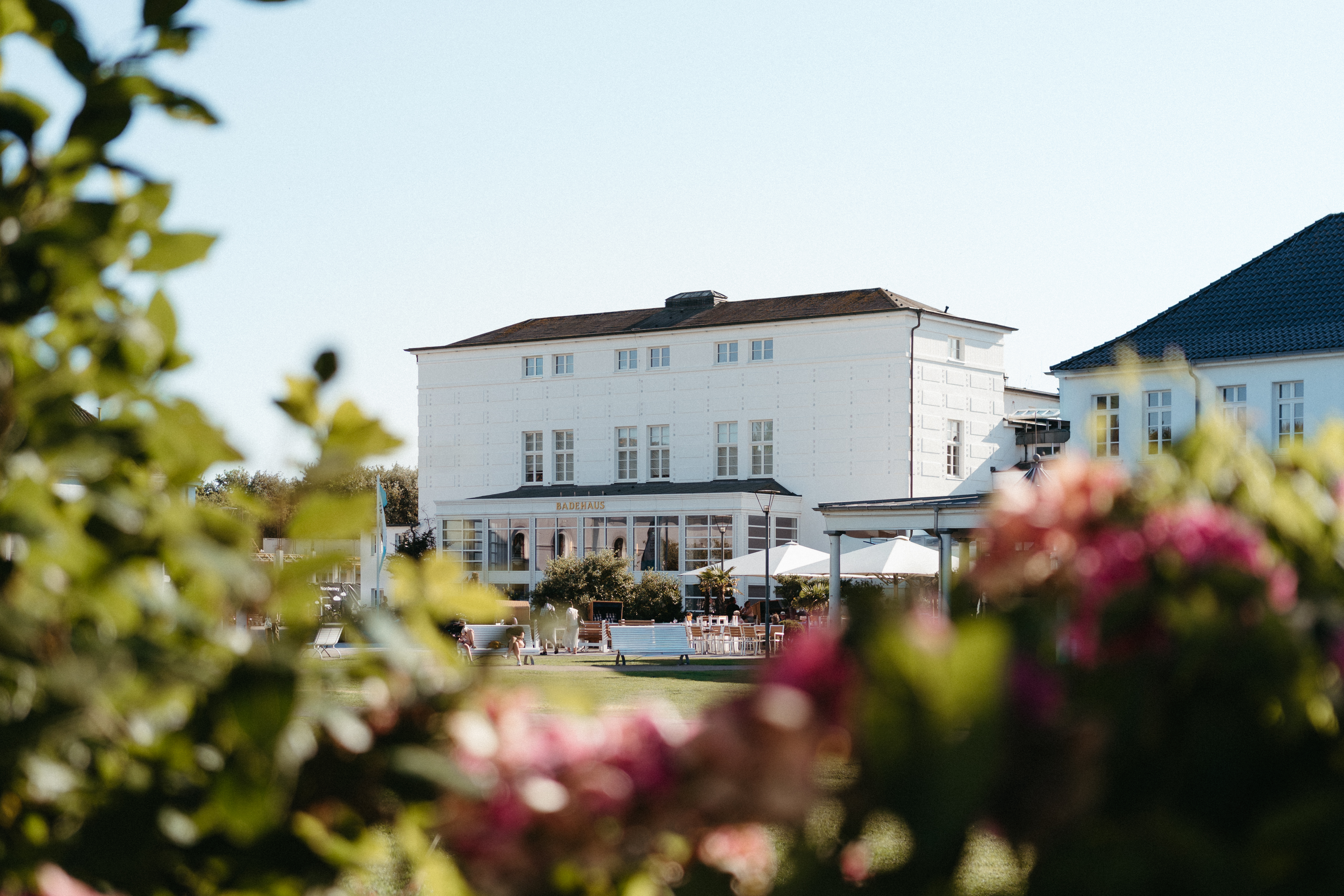 Weißes Wohnhaus mit Terrasse und Sonnenschirmen, im Vordergrund blühende Sträucher.