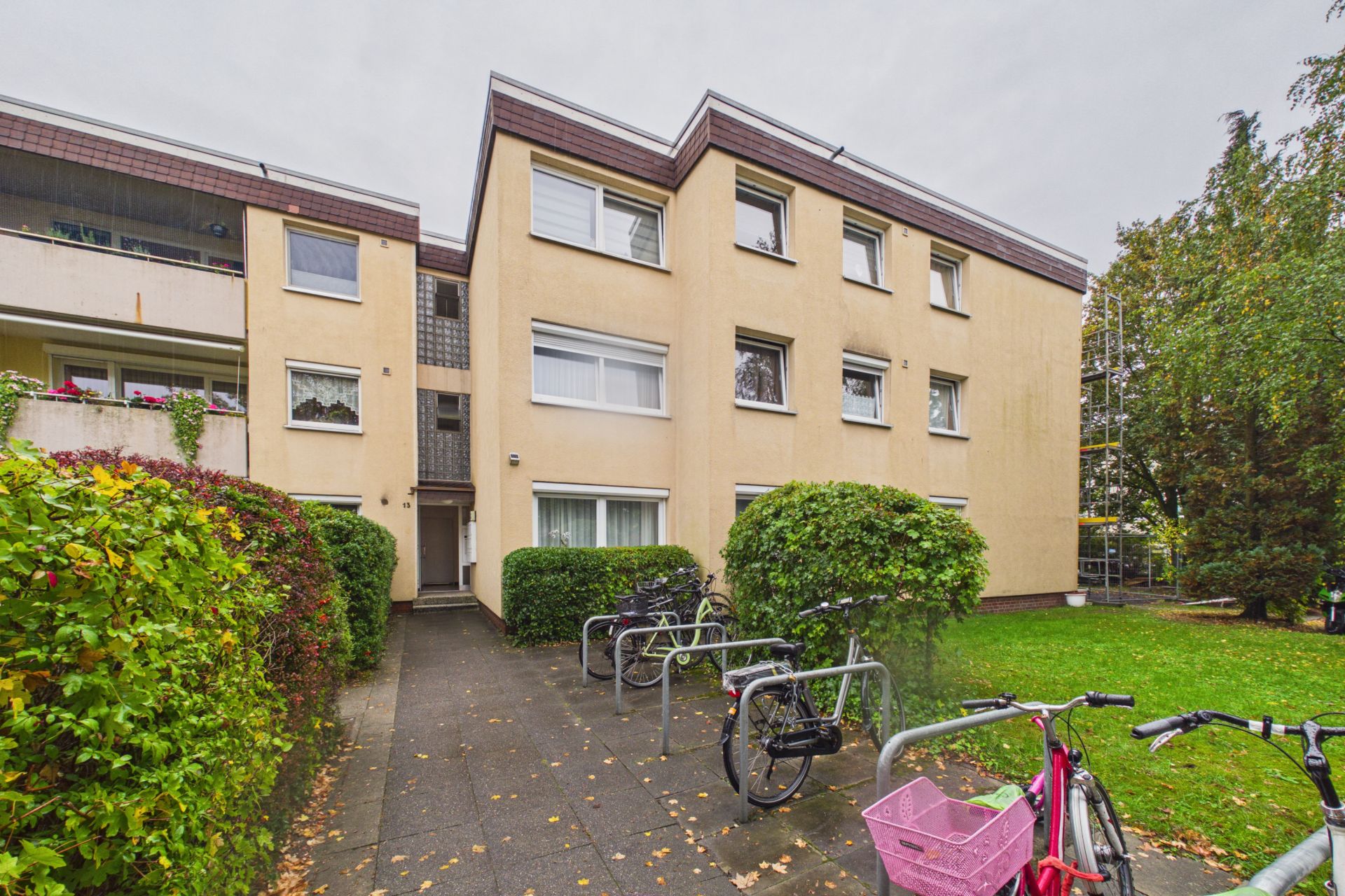 Mehrfamilienhaus mit drei Etagen, Fahrradbügeln und Büschen im Vorgarten.