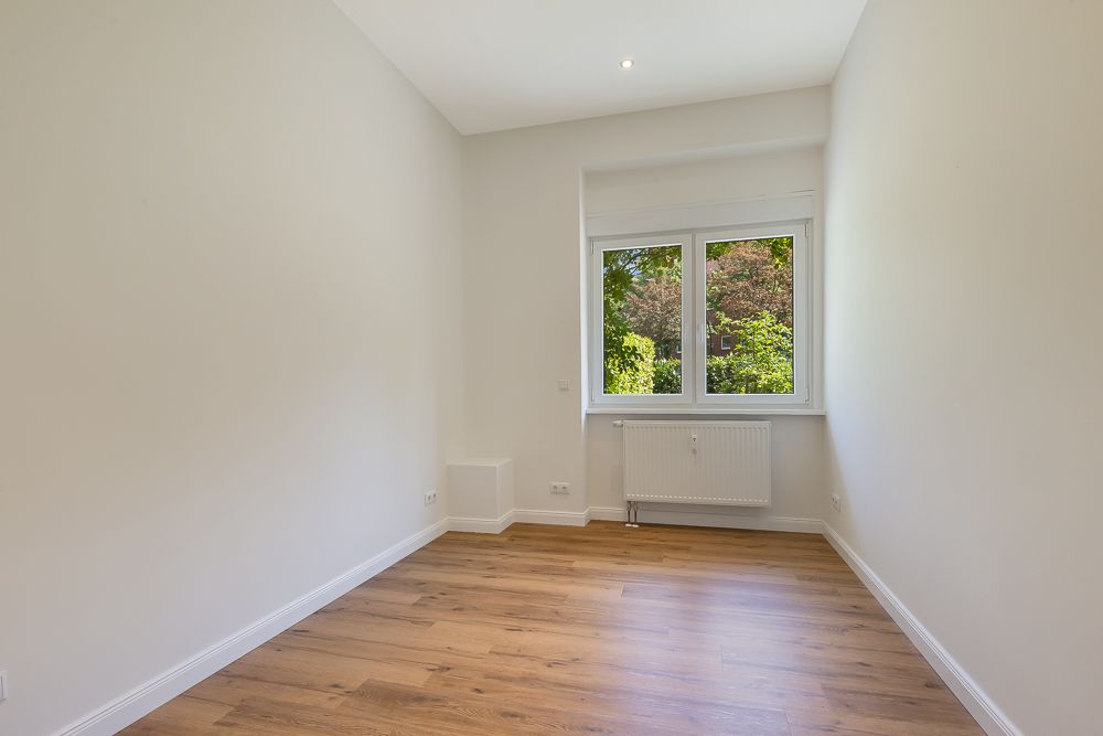 Leeres Zimmer mit weiß gestrichenen Wänden, Holzfußboden und Fenster mit Blick ins Grüne.