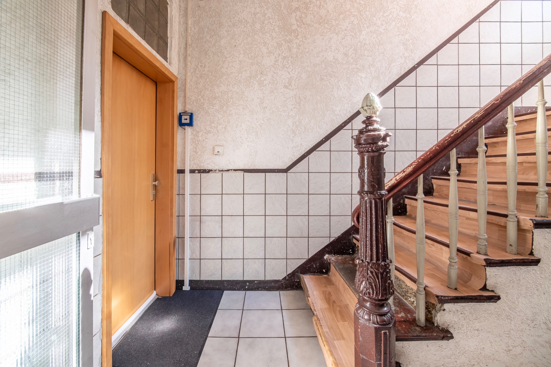 Treppenhaus mit Holztreppe, Kacheln an der Wand und einer braunen Tür auf der linken Seite.