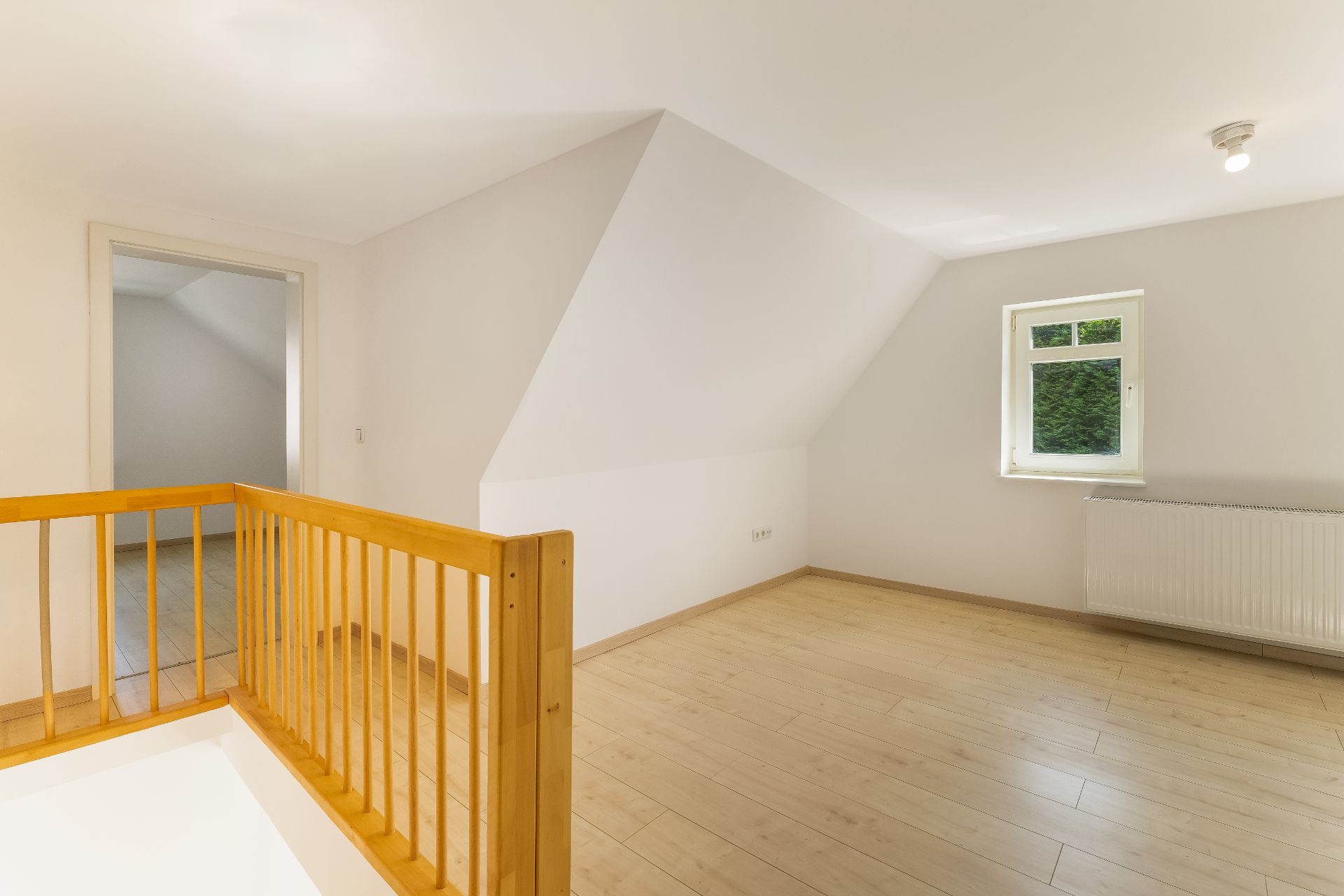 Helles Zimmer mit schräger Decke, Holzgeländer, Fenster und Heizkörper.
