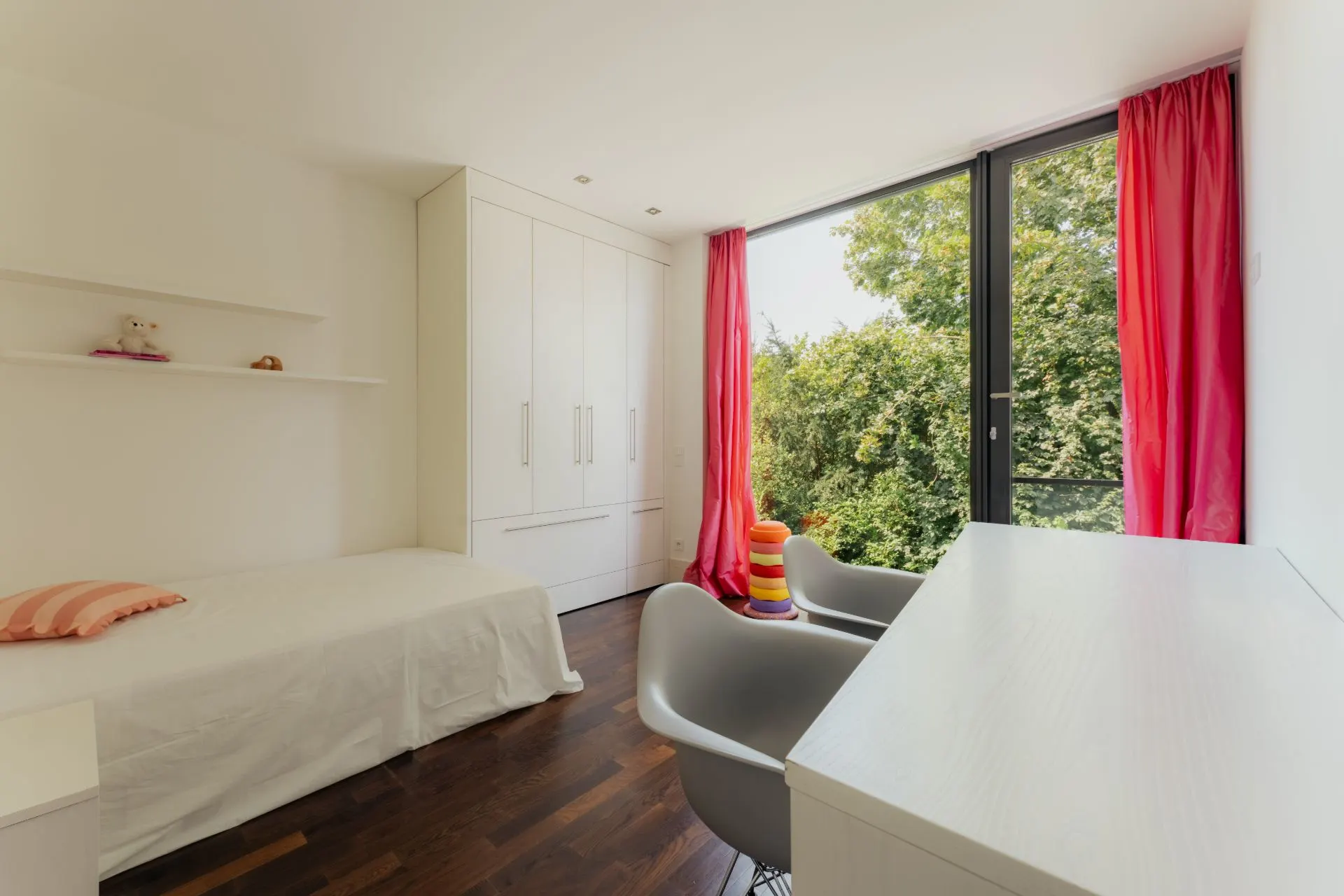 Helles Schlafzimmer mit Einzelbett, Schreibtisch, zwei Stühlen und rotem Vorhang vor großem Fenster.