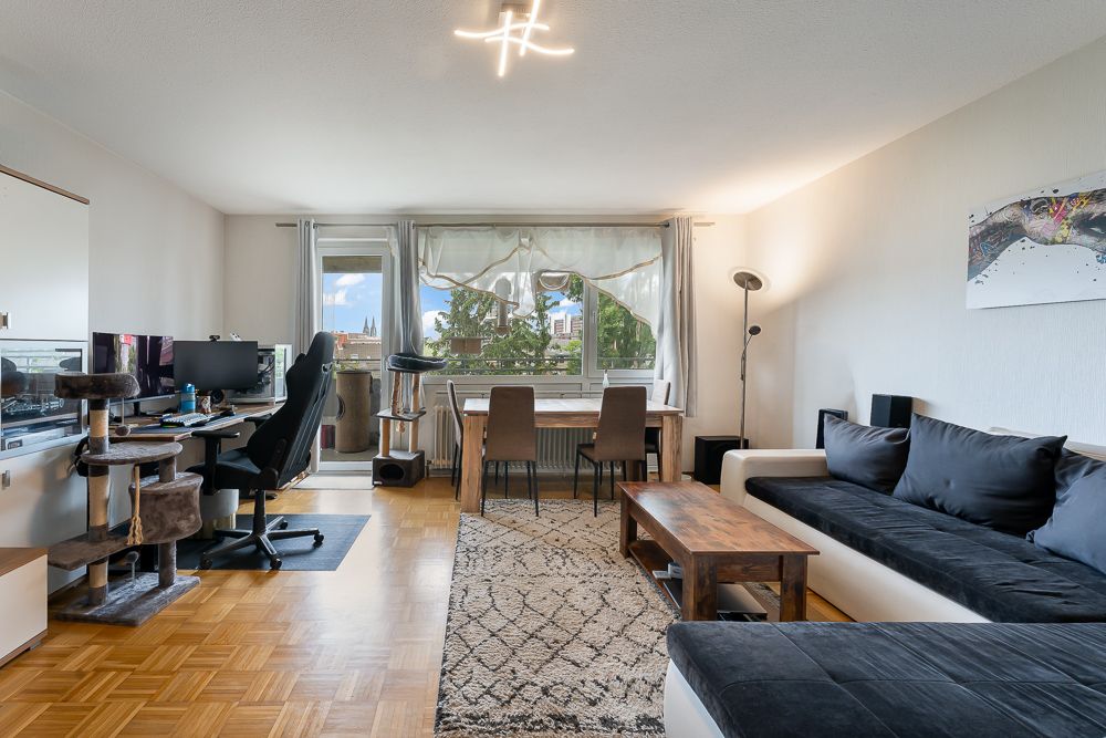 Helles Wohnzimmer mit Sofa, Schreibtisch, Esstisch, Teppich und großem Fenster.