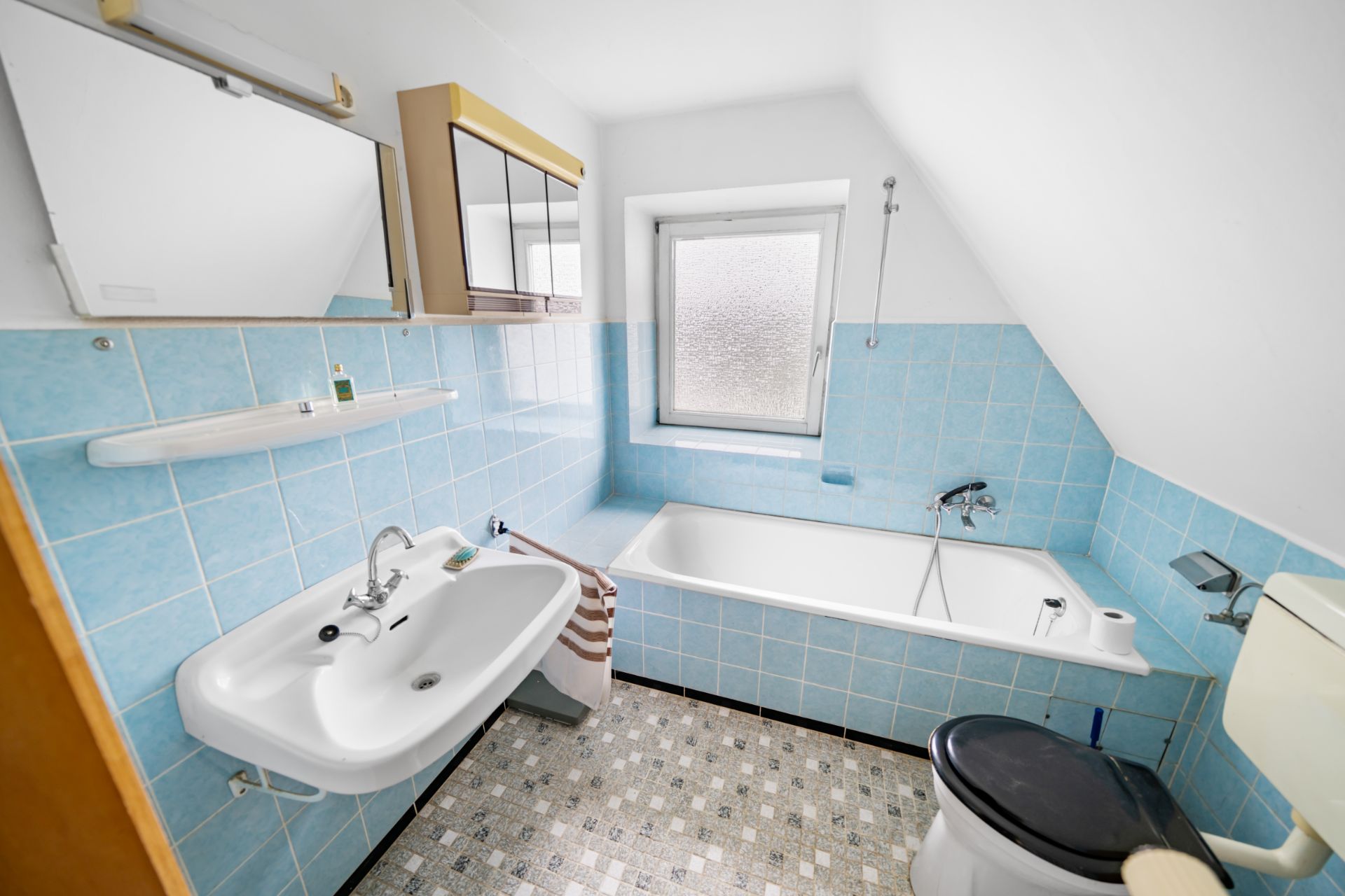 Kleines Badezimmer mit Badewanne, Waschbecken, Toilette und Fenster, blaue Fliesen an den Wänden.