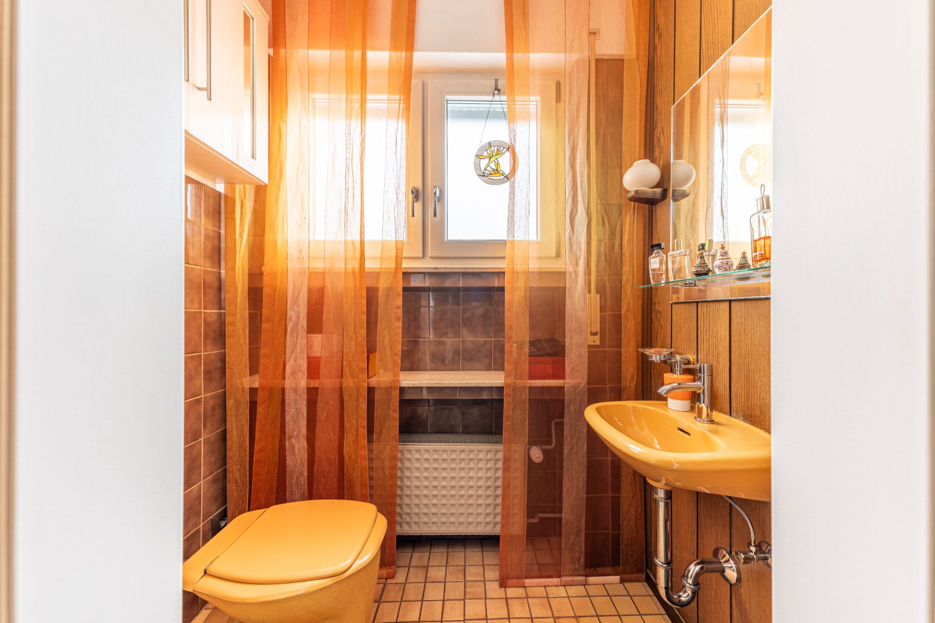 Kleines Bad mit gelber Toilette, Waschbecken, Spiegel und orangefarbenem Vorhang vor Fenster.