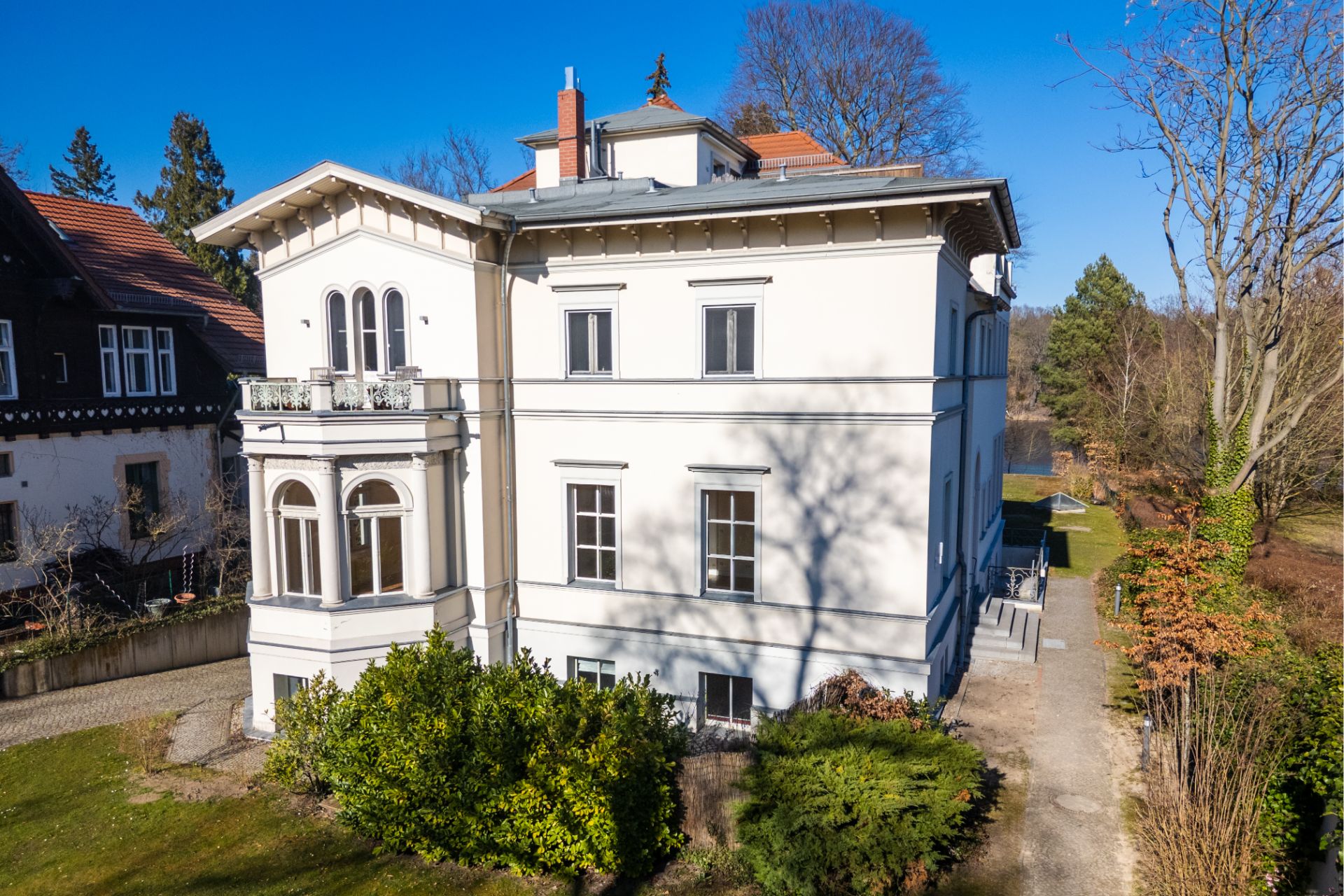 Großes, helles Wohnhaus mit mehreren Fenstern, Balkon und Garten, umgeben von Bäumen.