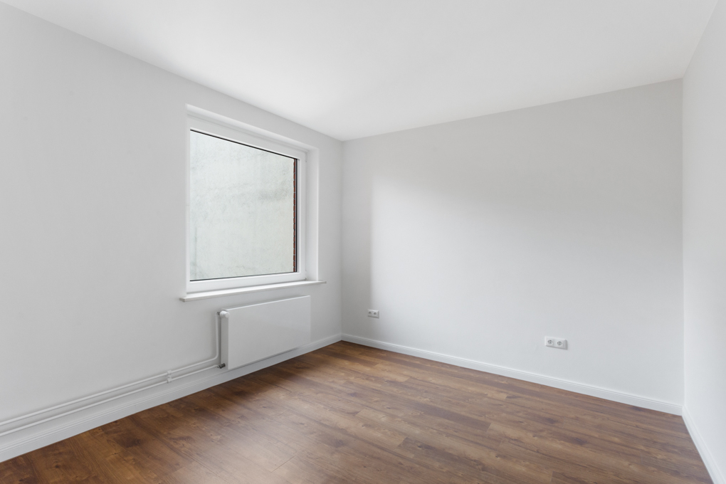 Leerer Raum mit weißen Wänden, großem Fenster und braunem Holzboden.