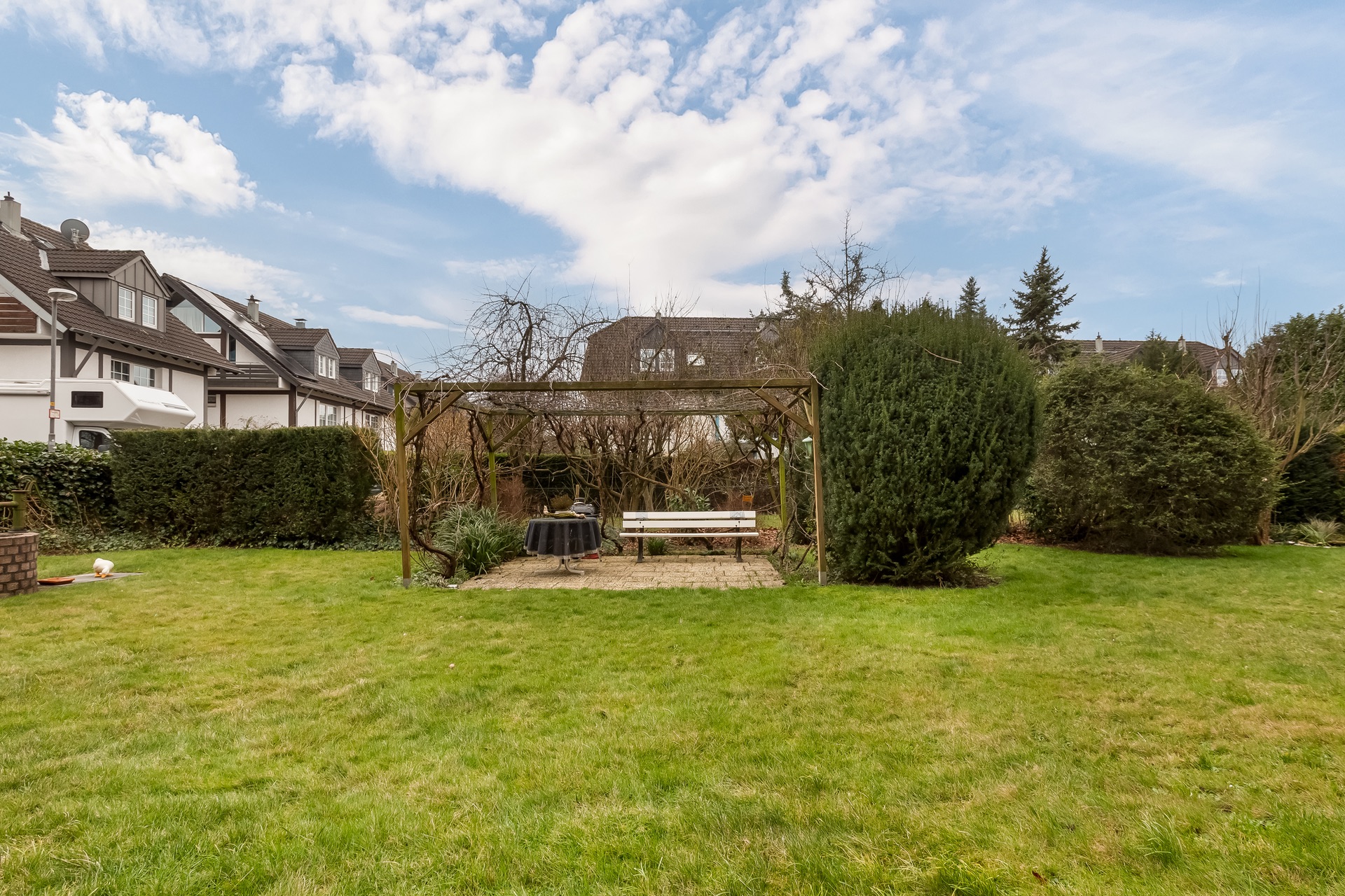 Garten mit Rasenfläche, Pergola, Bank und Tisch, umgeben von Sträuchern und Bäumen.