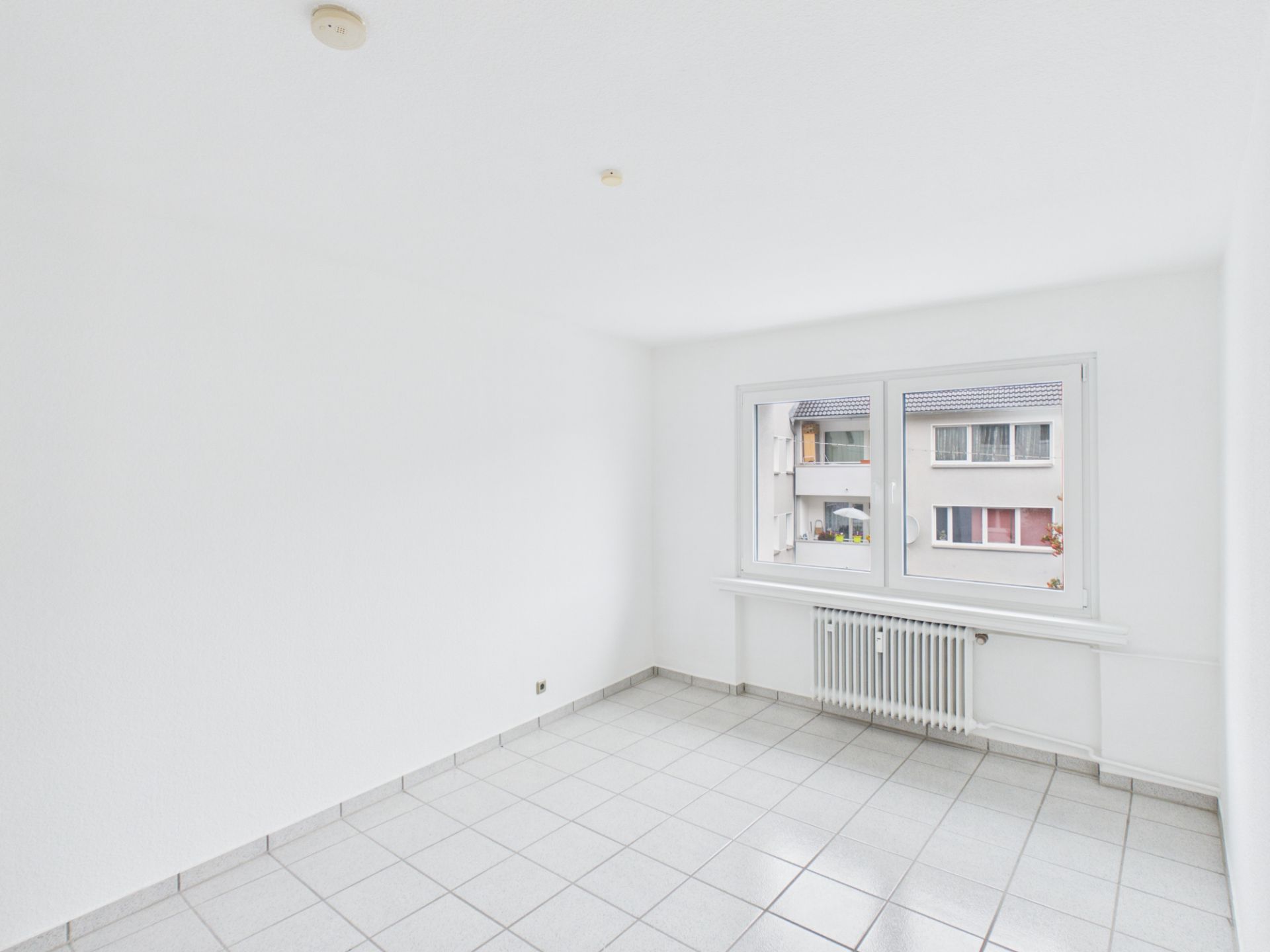 Leerer, heller Raum mit weißen Wänden, Fliesenboden und großem Fenster mit Heizkörper darunter.