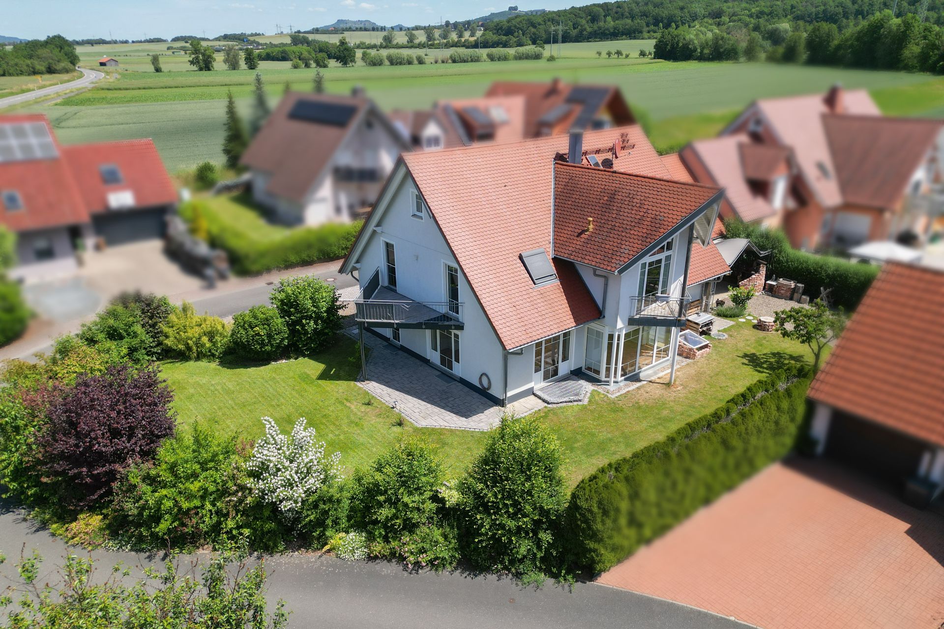 Modernes Einfamilienhaus mit rotem Dach und Garten, umgeben von Nachbarhäusern und Feldern.