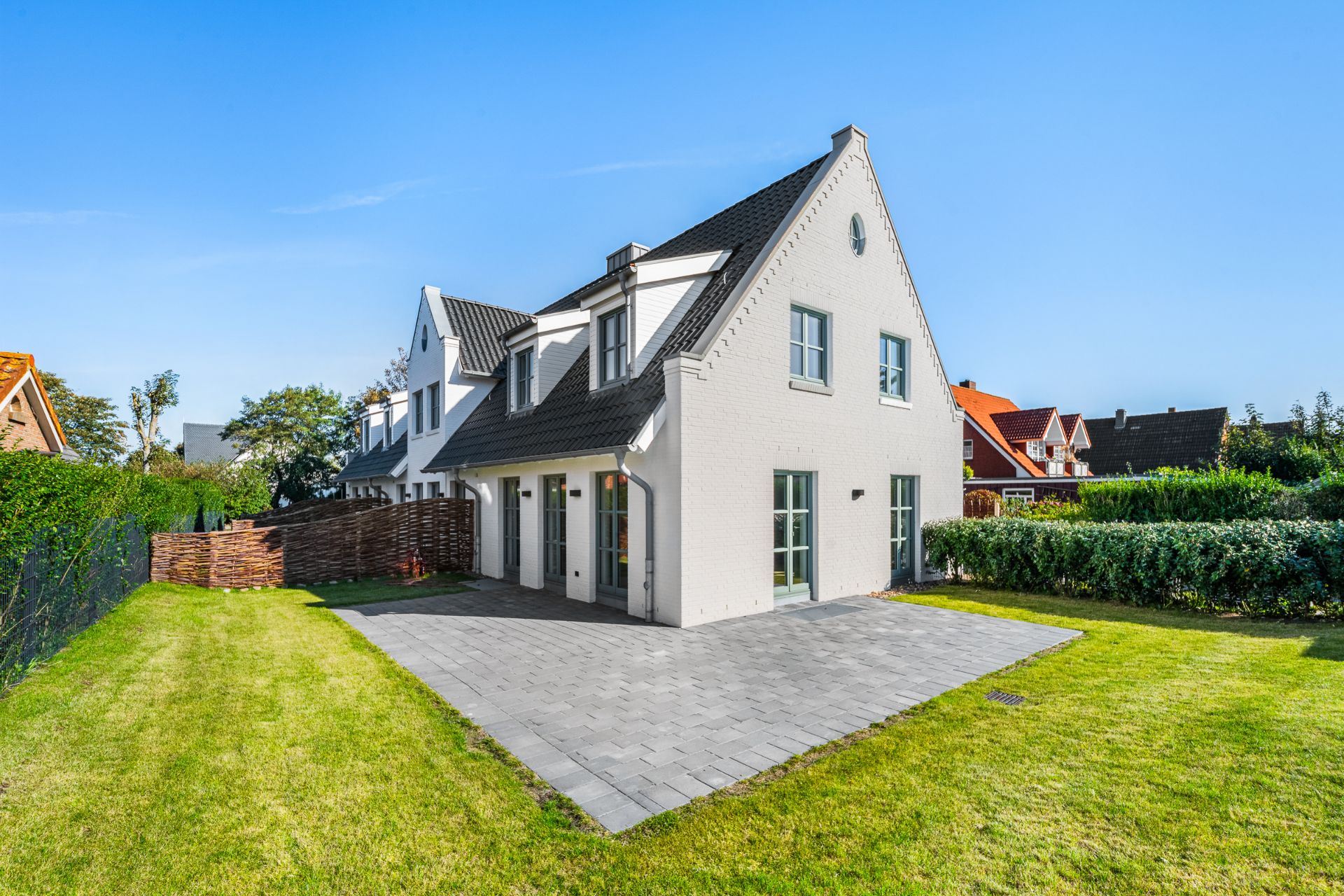Weißes Einfamilienhaus mit dunklem Dach, Terrasse und gepflegtem Garten bei sonnigem Wetter.