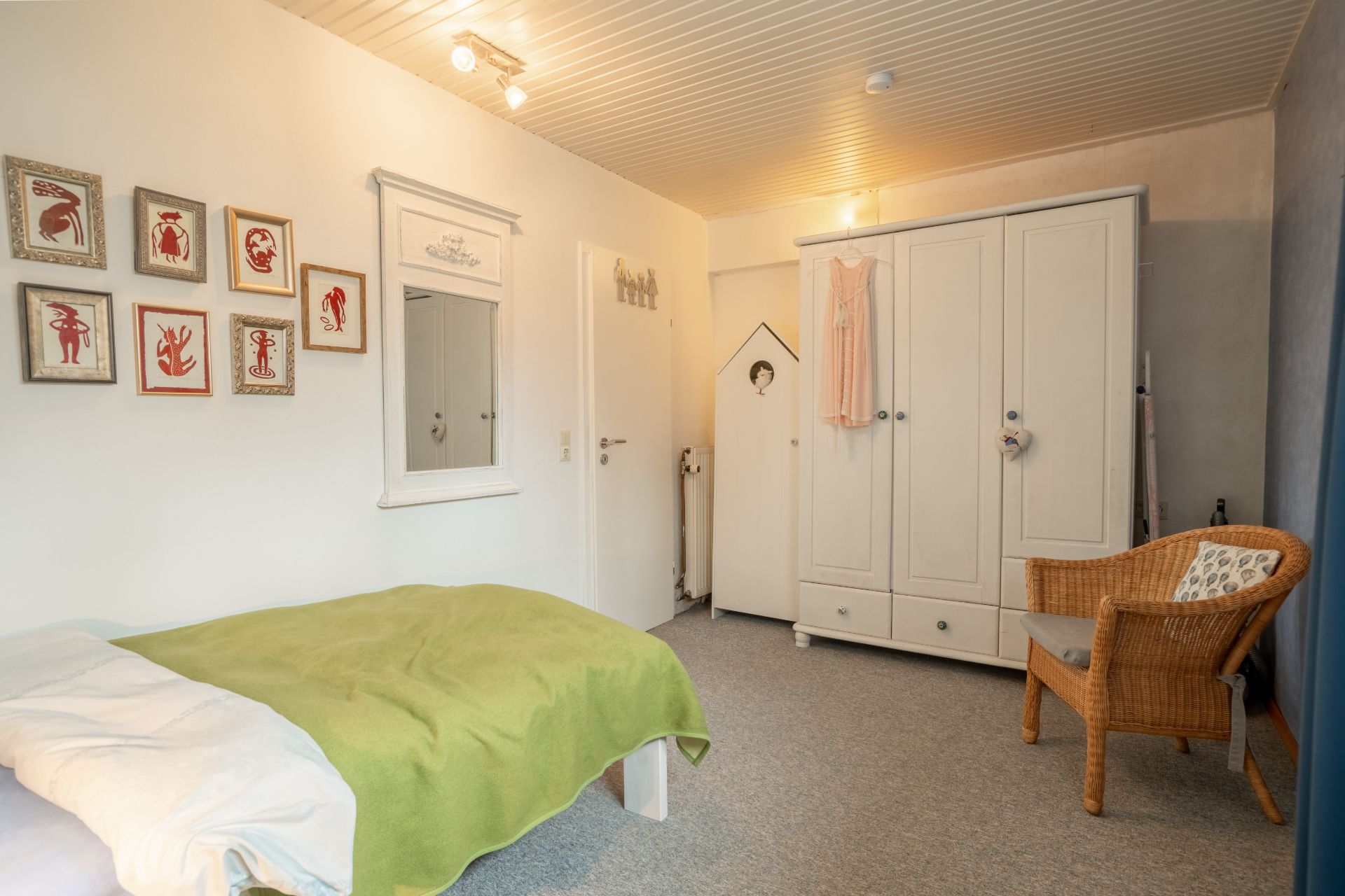 Helles Schlafzimmer mit Einzelbett, grünem Bettbezug, weißem Schrank und Stuhl aus Korbgeflecht.