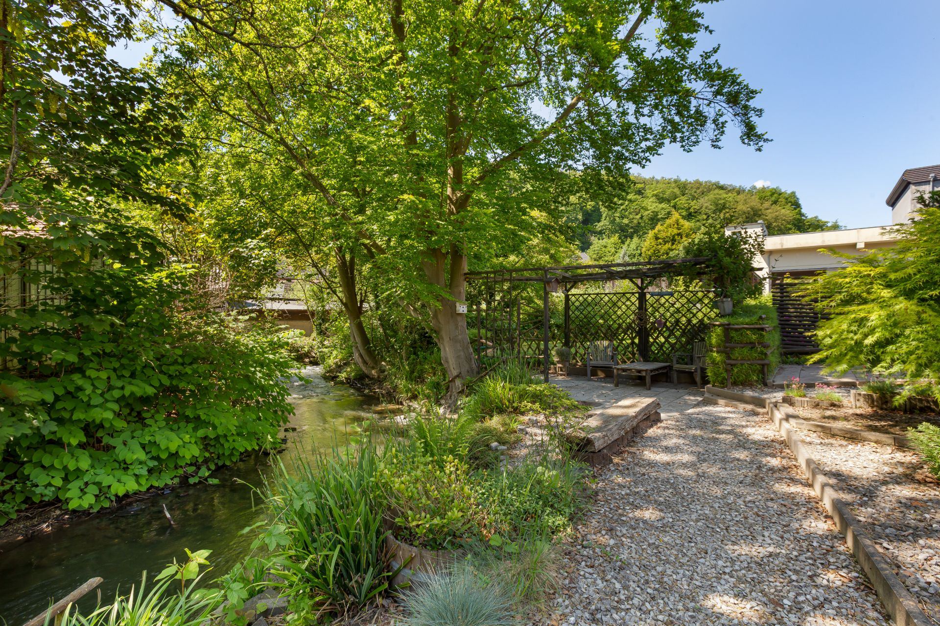 Garten mit Kiesweg, Bachlauf, viel Grün und einer Holzpergola mit Sitzbank.