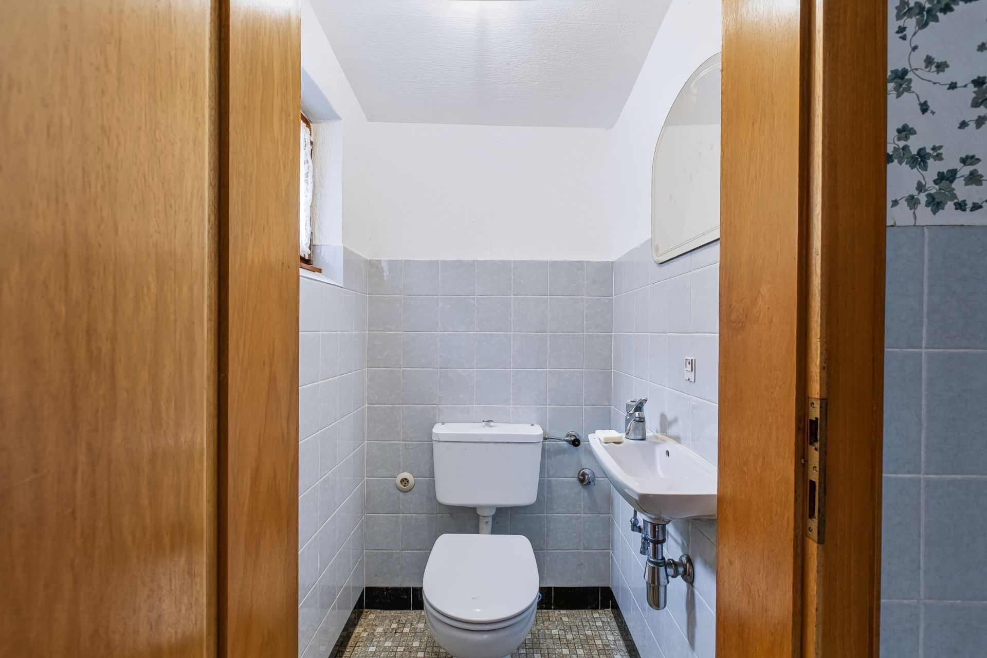 Kleines Badezimmer mit Toilette, Waschbecken und Spiegel, Wandfliesen und kleinem Fenster.