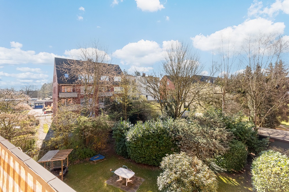 Blick vom Balkon auf einen Garten mit Bäumen, Sträuchern, kleinem Spielplatz und umliegende Wohnhäuser.