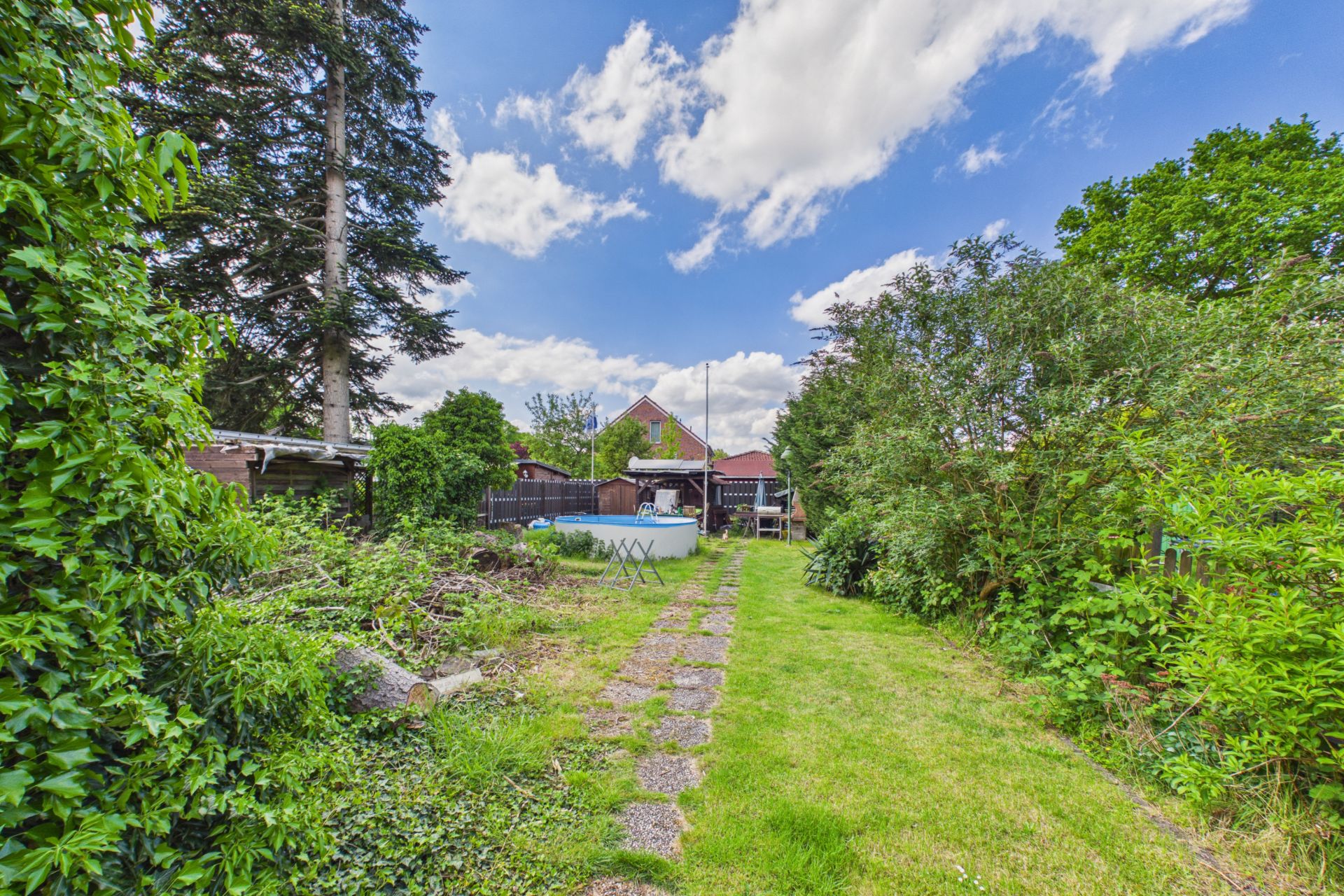 Garten mit Rasen, kleinem Pool und Gartenweg, umgeben von Bäumen und Sträuchern, Haus im Hintergrund.