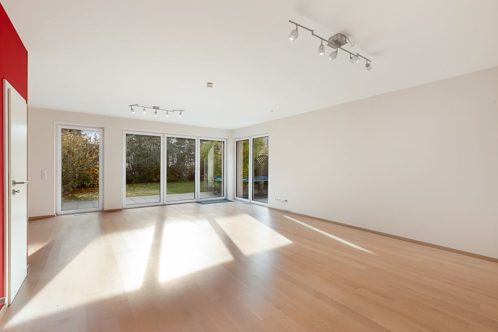 Heller, leerer Wohnraum mit Holzfußboden und großen Fenstern zum Garten.
