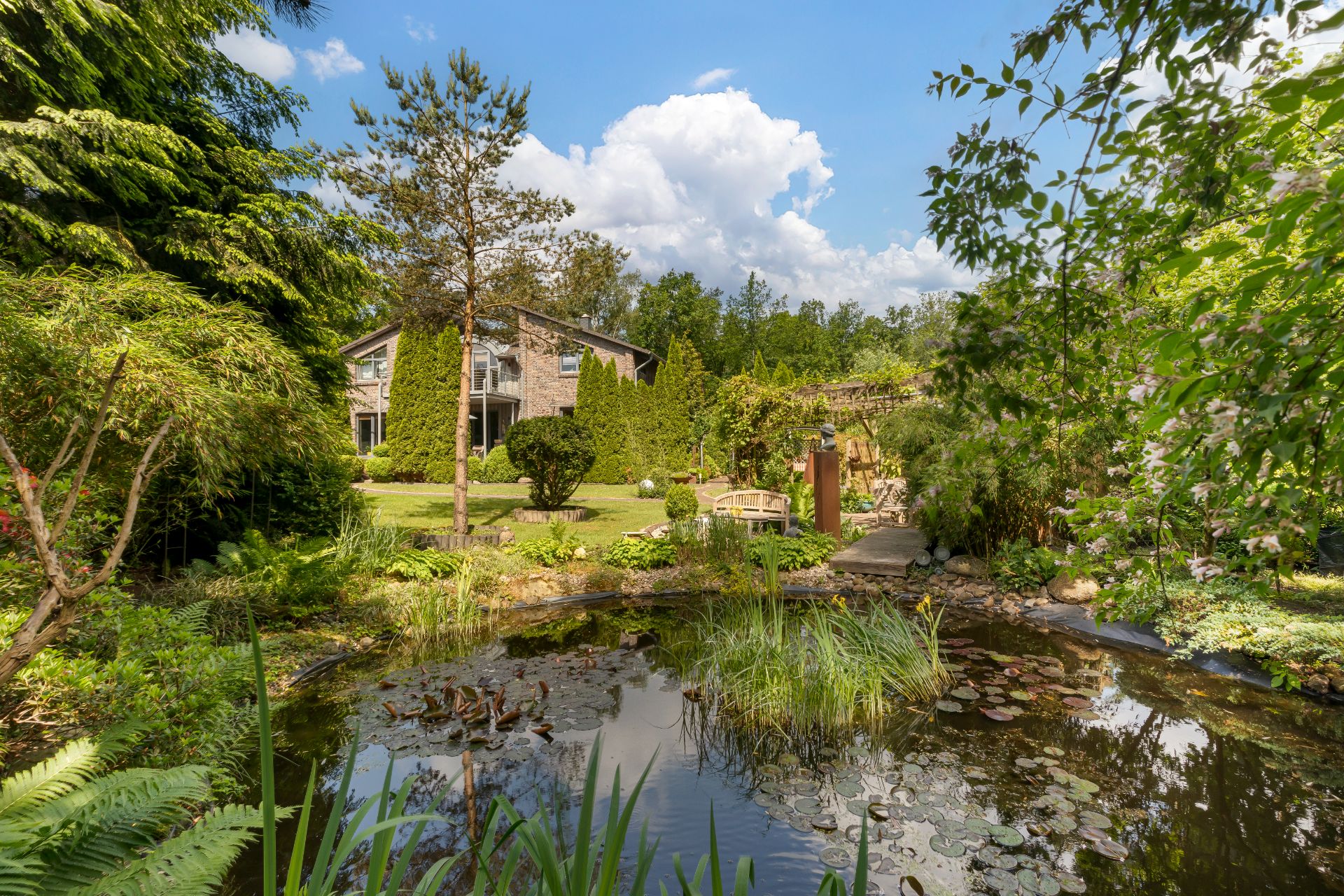 Garten mit Teich, Sitzbank und viel Grün vor einem modernen Wohnhaus an einem sonnigen Tag.