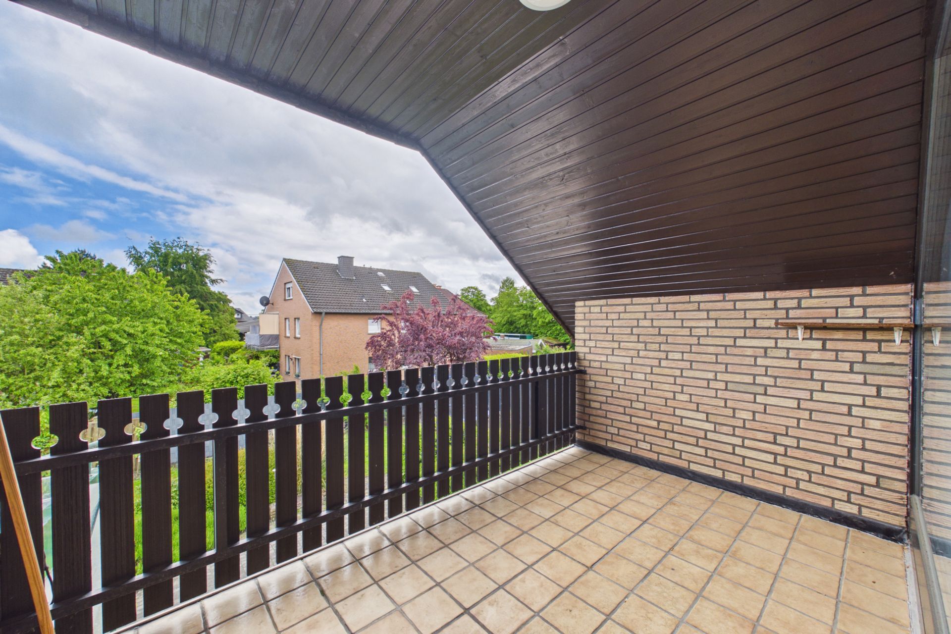 Überdachter Balkon mit Fliesenboden und Holzzaun, Blick auf Nachbarhäuser und Bäume.