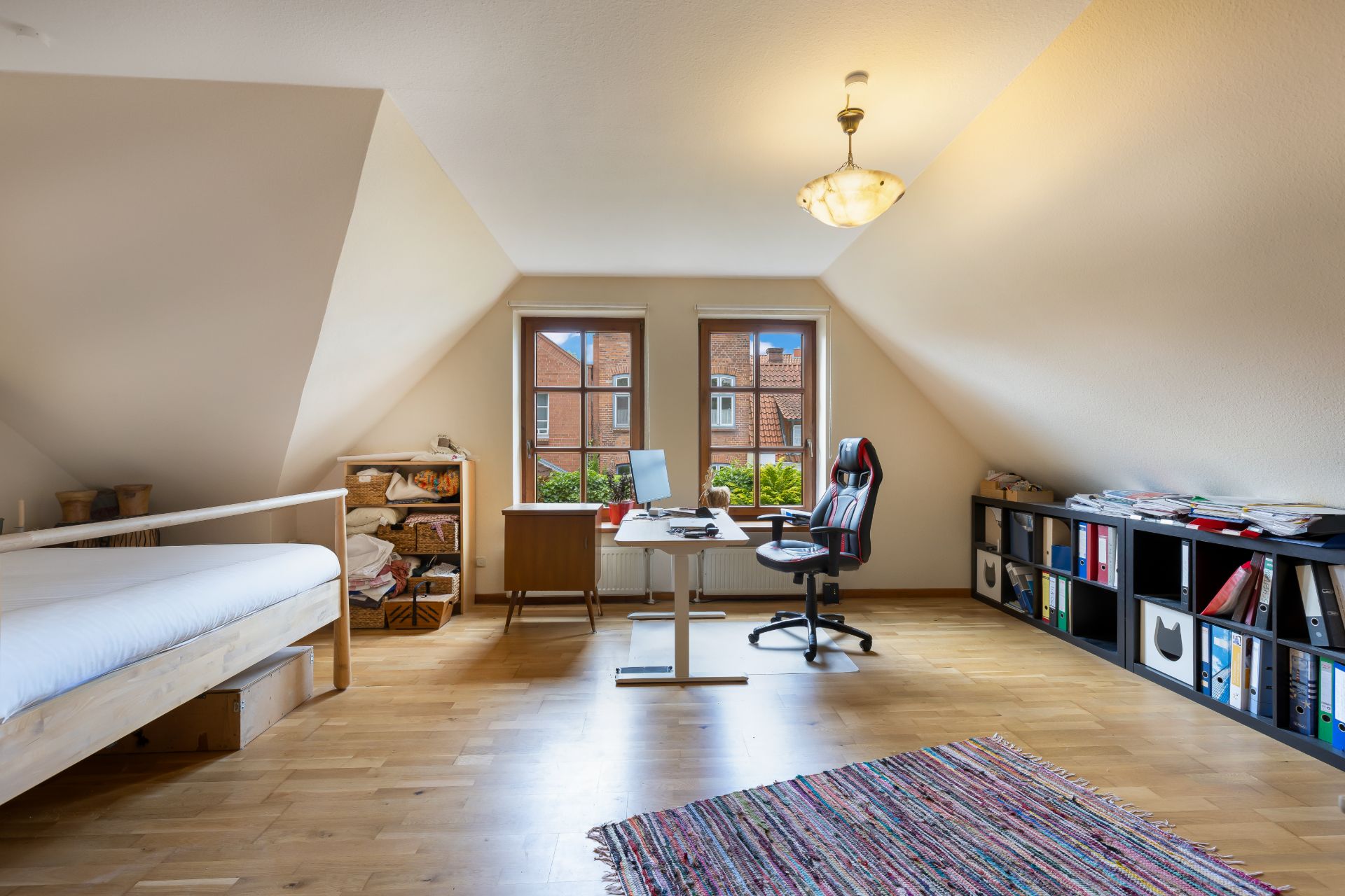 Helles Zimmer mit Schreibtisch, Stuhl, Regal, Bett und zwei Fenstern, Holzboden und Teppich.