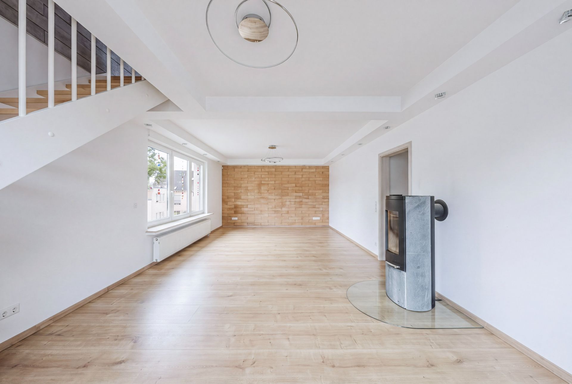 Helles Wohnzimmer mit Holzfußboden, Kamin und Treppe, große Fenster an der linken Wand.