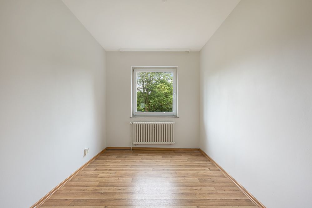 Leerer Raum mit Holzfußboden, weiß gestrichenen Wänden und Fenster mit Blick ins Grüne.