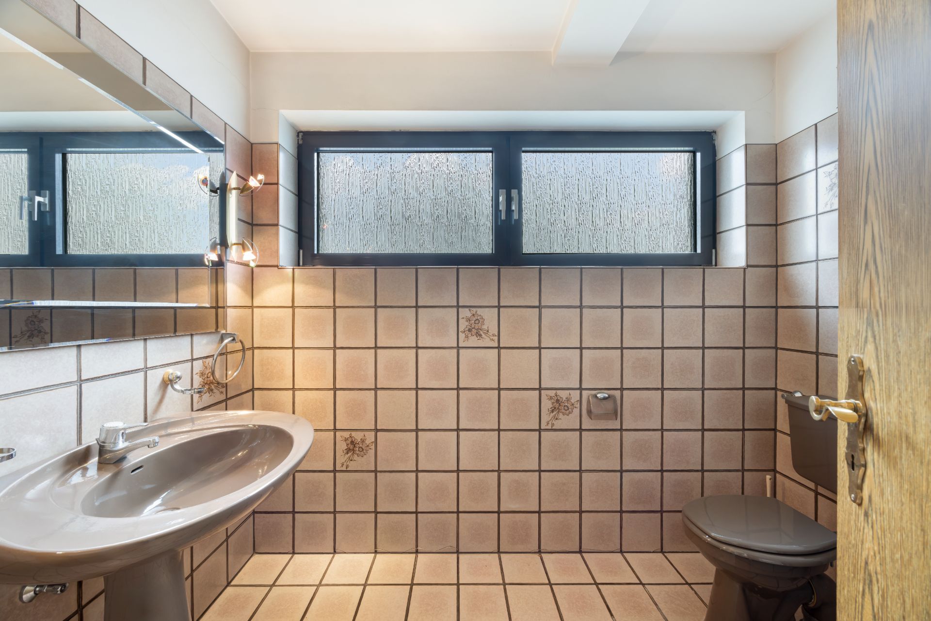 Badezimmer mit braunen Fliesen, Waschbecken, Spiegel, Fenster und Toilette.