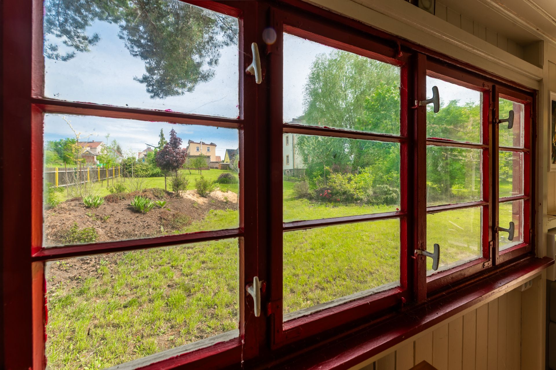 Blick durch ein großes rotes Fenster auf einen grünen Garten mit Bäumen und Rasen.