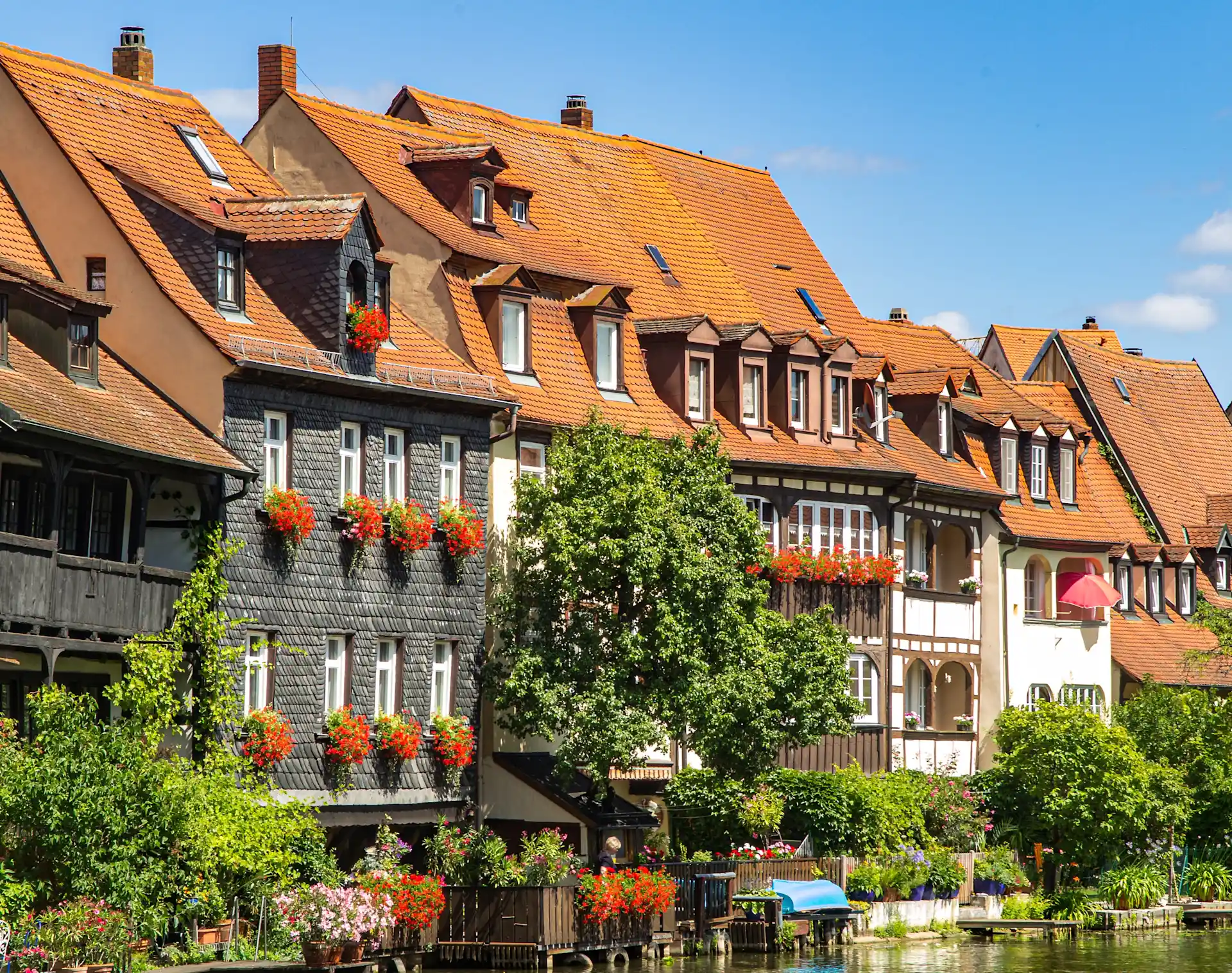 Mehrere historische Wohnhäuser mit roten Dächern und Blumenkästen an den Fenstern am Flussufer.