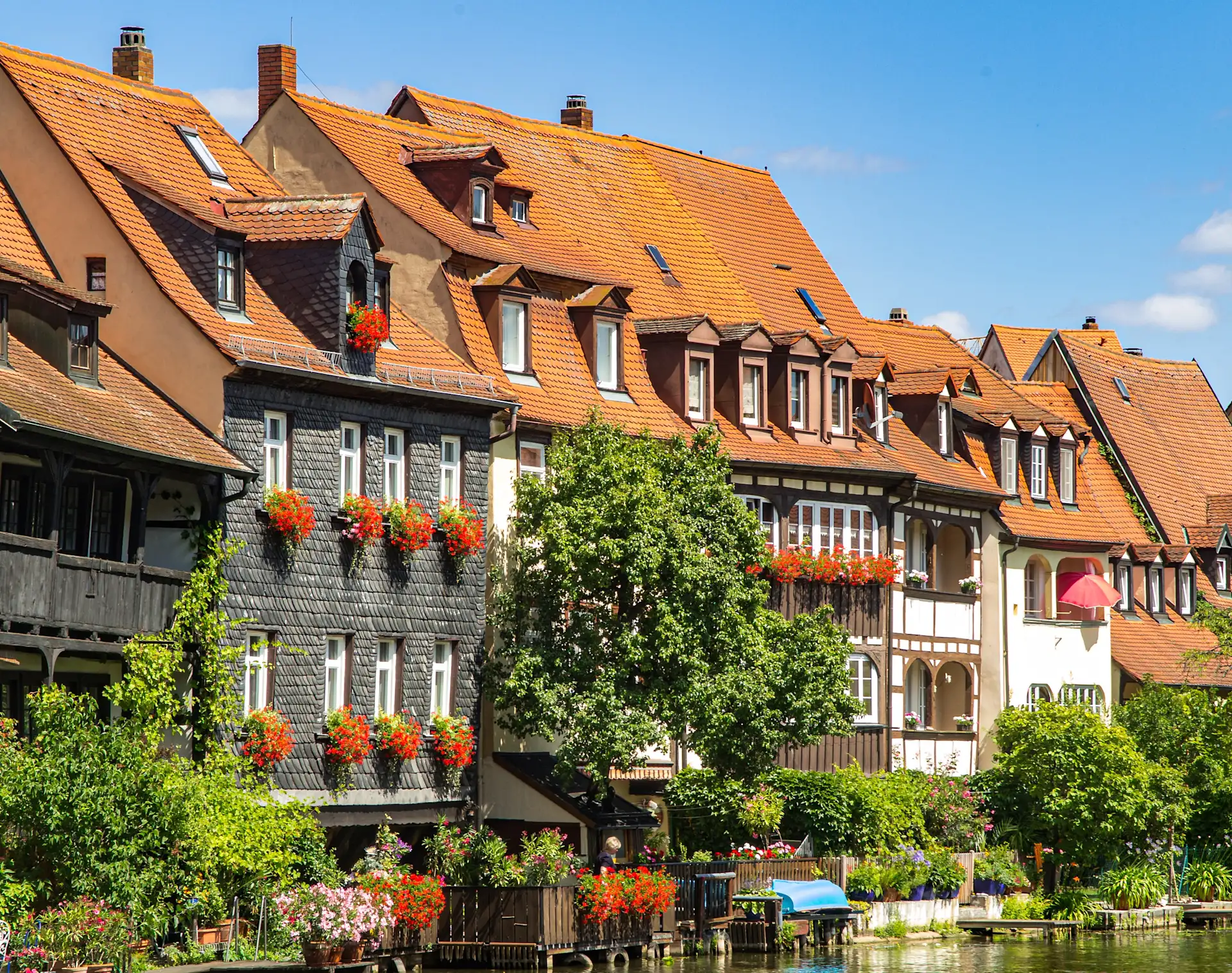 Mehrere historische Wohnhäuser mit roten Dächern und Blumenkästen an den Fenstern am Flussufer.